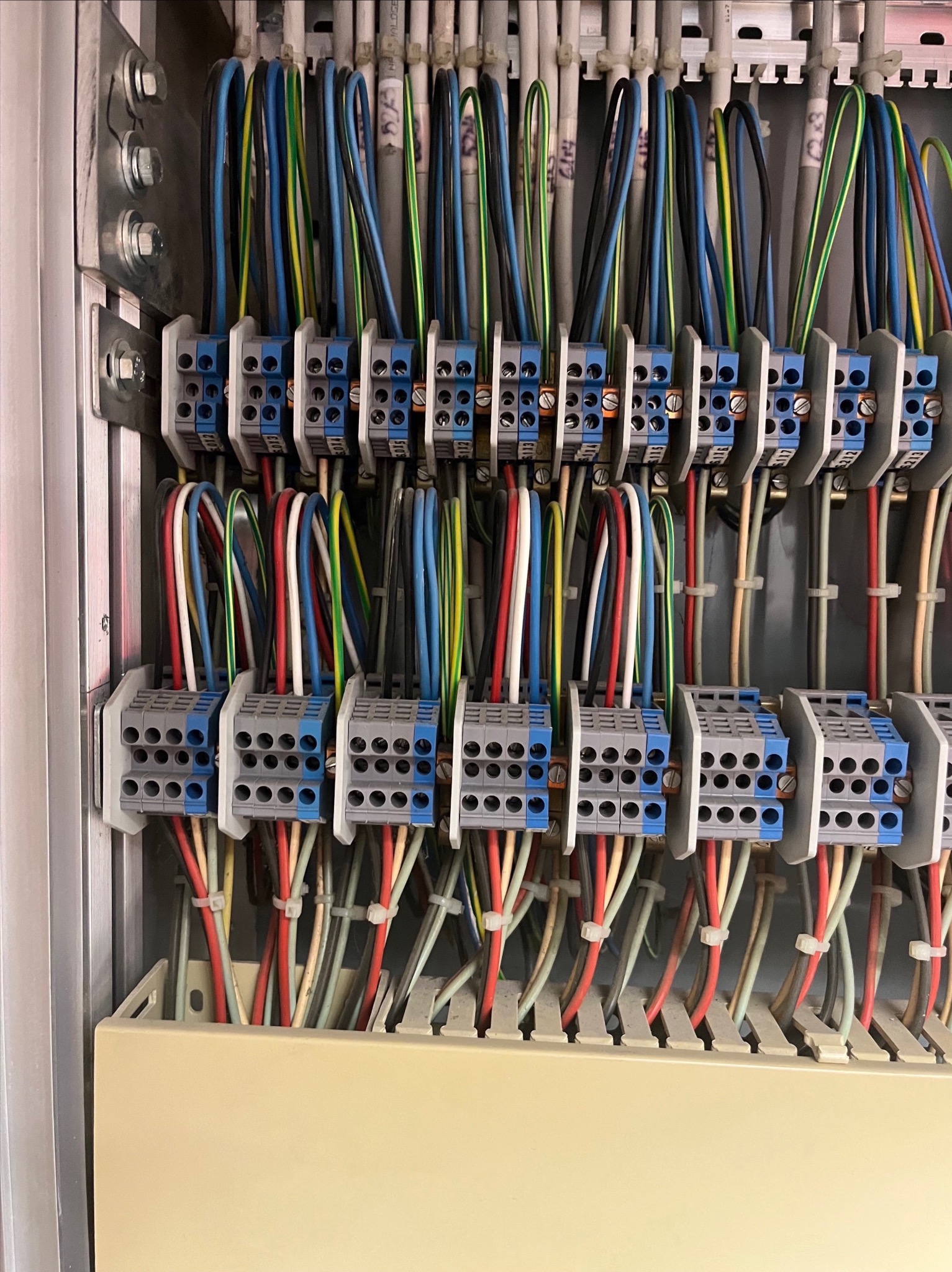 Rozdzielnica elektryczna z uporządkowanymi przewodami o różnych kolorach, podłączonymi do listew zaciskowych z oznaczeniami na górze, widoczne elementy montażowe na metalowej obudowie.