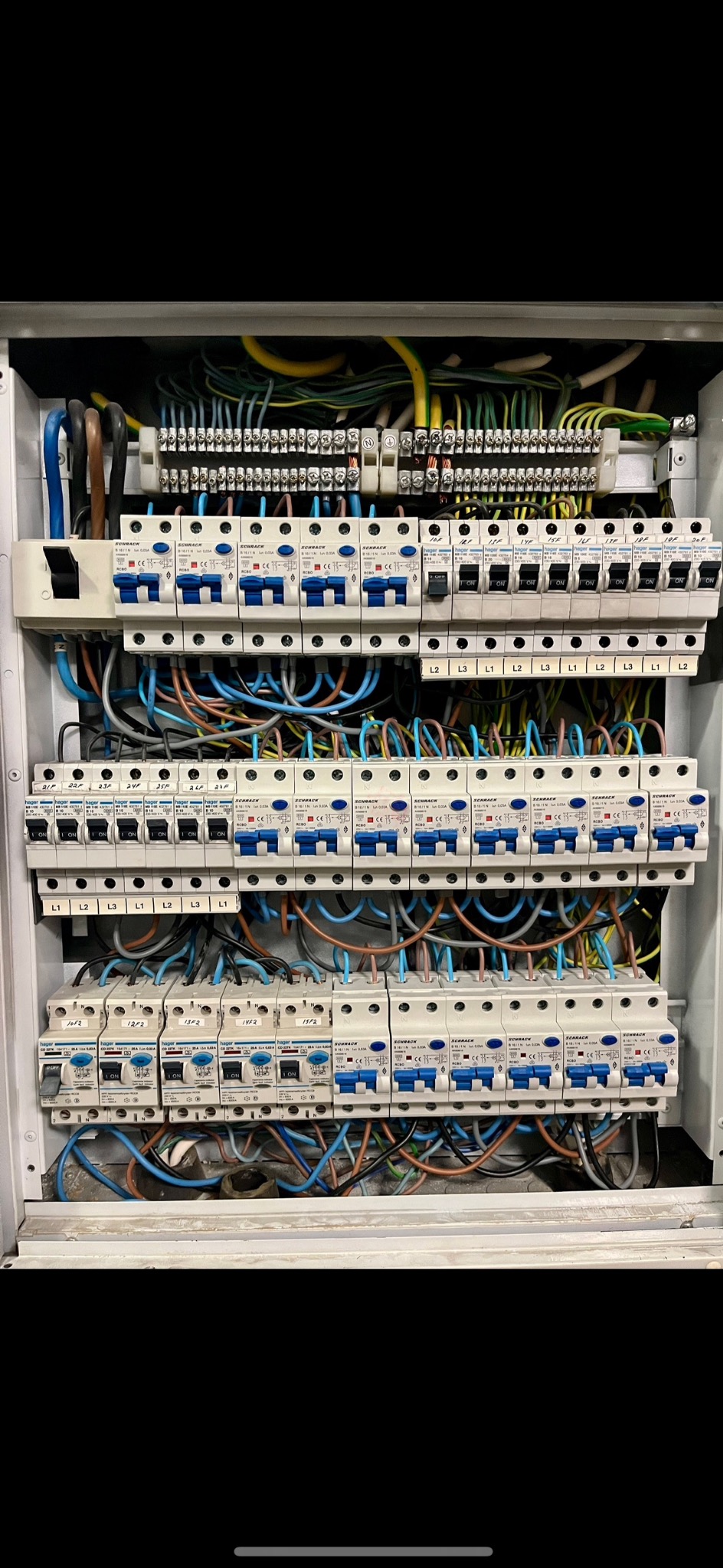 Rozdzielnia elektryczna z licznymi bezpiecznikami i przewodami w różnych kolorach, zamontowana w szarej obudowie. Widoczne oznaczenia L1, L2, L3 oraz napisy na bezpiecznikach.