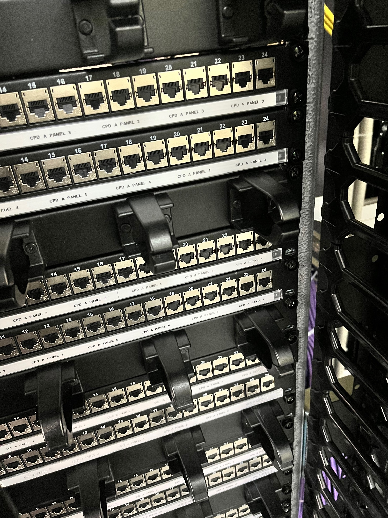 Panel krosowniczy z portami RJ45 w szafie rack, widoczne numery portów i oznaczenia paneli, czarne uchwyty kablowe, oświetlenie studyjne podkreśla detale.