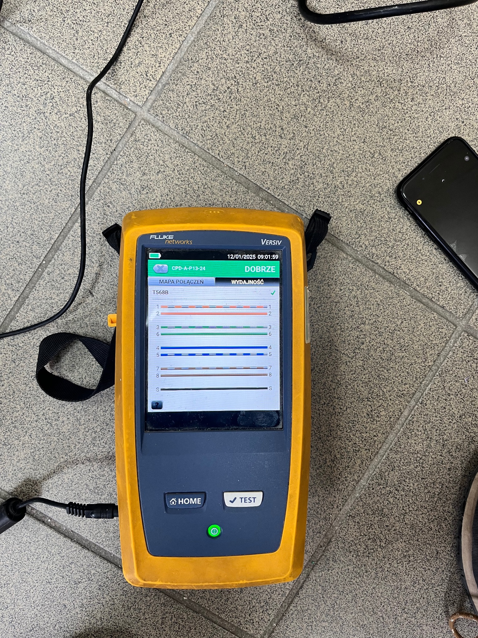 Tester okablowania Fluke Networks Versiv z pozytywnym wynikiem testu mapy połączeń T568B, leżący na szarej podłodze z kafelków. Widoczny fragment kabla i smartfona.