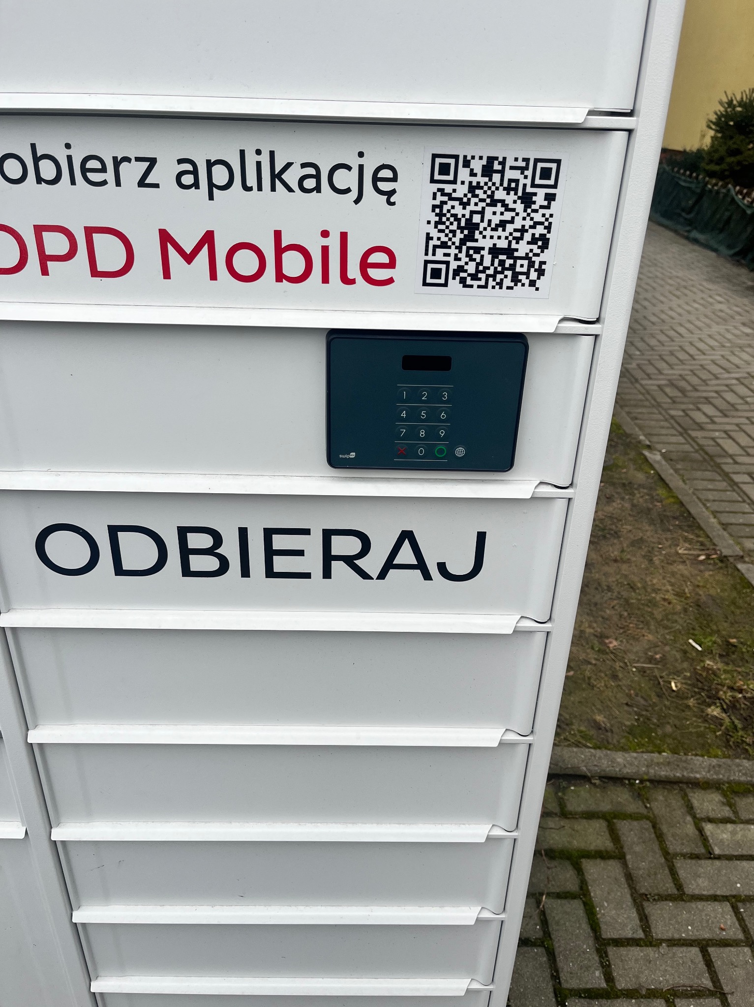 Panel sterowania paczkomatu z klawiaturą numeryczną i napisem 'ODBIERAJ'. Widoczny kod QR do pobrania aplikacji mobilnej. Ujęcie z dołu, na pierwszym planie chodnik.
