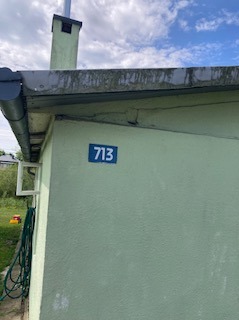 Fragment elewacji budynku z numerem 713, widoczny komin i rynna, jasna kolorystyka, niebieska tabliczka z numerem.