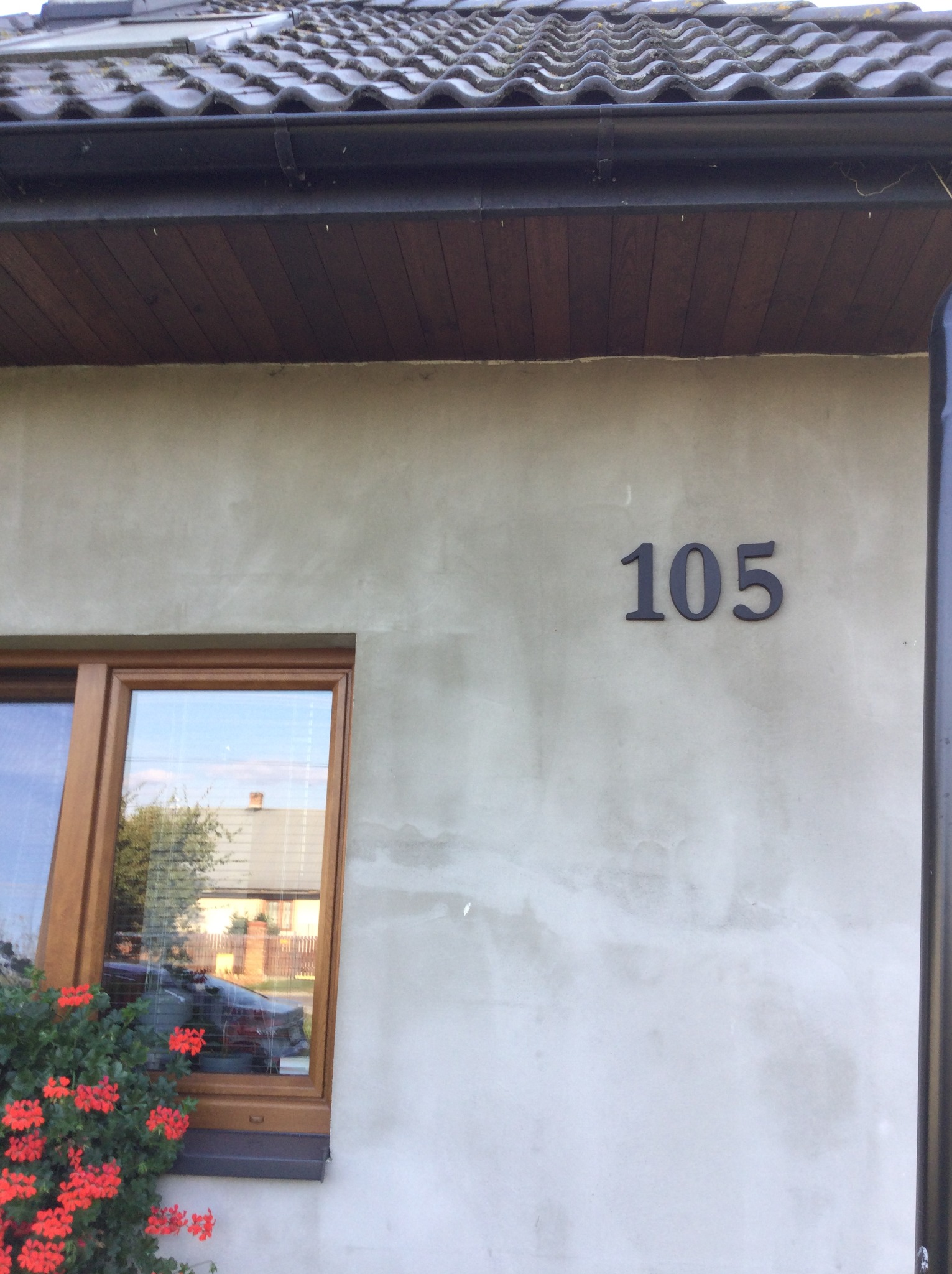 Elewacja budynku z numerem 105, okno z drewnianą ramą i czerwonymi kwiatami w doniczce, widoczne fragmenty dachu i rynny.