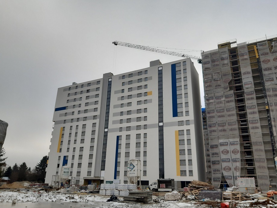 Wielopiętrowy budynek w trakcie budowy, widoczny żuraw budowlany i materiały budowlane na placu, drugi budynek obłożony folią ochronną.