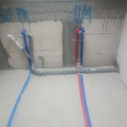 HydroDamps - Szare i kolorowe rury instalacji wodnej zamocowane do ściany z betonu komórkowego. Widoczne fragmenty niebieskich i czerwonych rur. Ślady po montażu w ścianie.