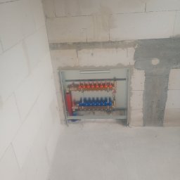 HydroDamps - Rozdzielacz ogrzewania podłogowego w szarej obudowie, zamontowany na ścianie z betonu komórkowego. Widoczne czerwone i niebieskie zawory.