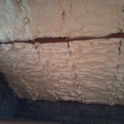 Tomasz Grajner - Poddasze z widoczną warstwą izolacji termicznej z pianki poliuretanowej, przymocowanej do krokwi za pomocą metalowych klamer, oraz fragmentem muru z ciemnych cegieł.