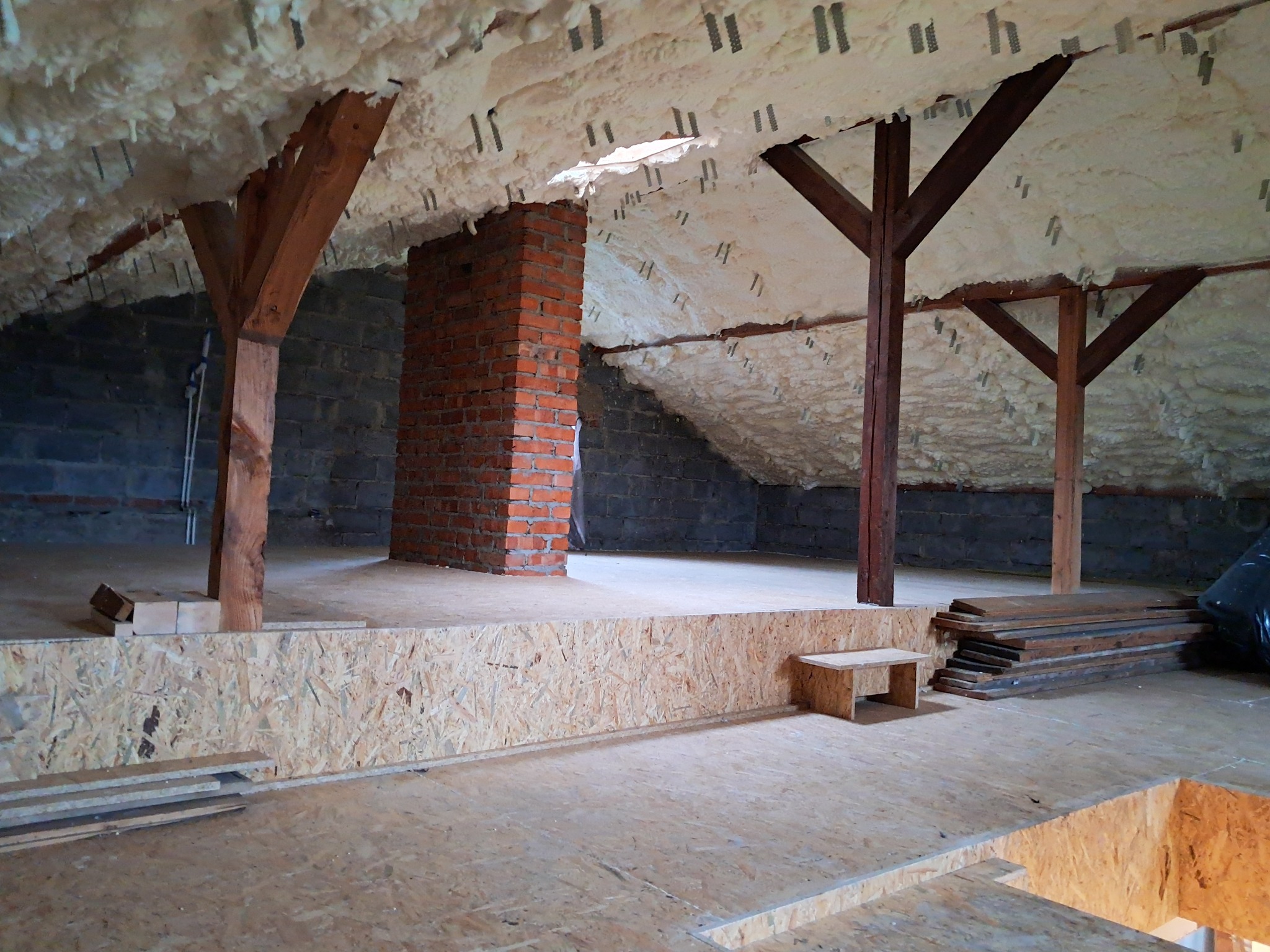 Poddasze z widoczną izolacją piankową, drewnianymi belkami i fragmentem komina z czerwonej cegły, podłoga wyłożona płytami OSB.