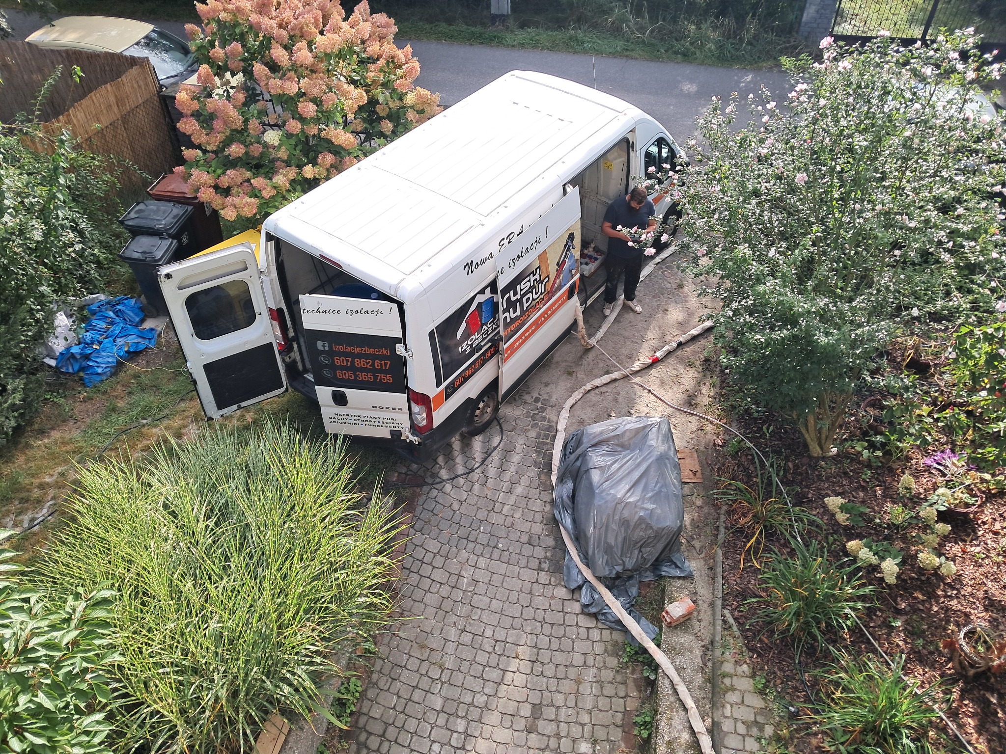 Widok z góry na biały samochód dostawczy z otwartymi drzwiami, z którego pracownik przygotowuje się do natrysku pianki izolacyjnej; na samochodzie widoczne reklamy firmy i numery telefonów, obok...