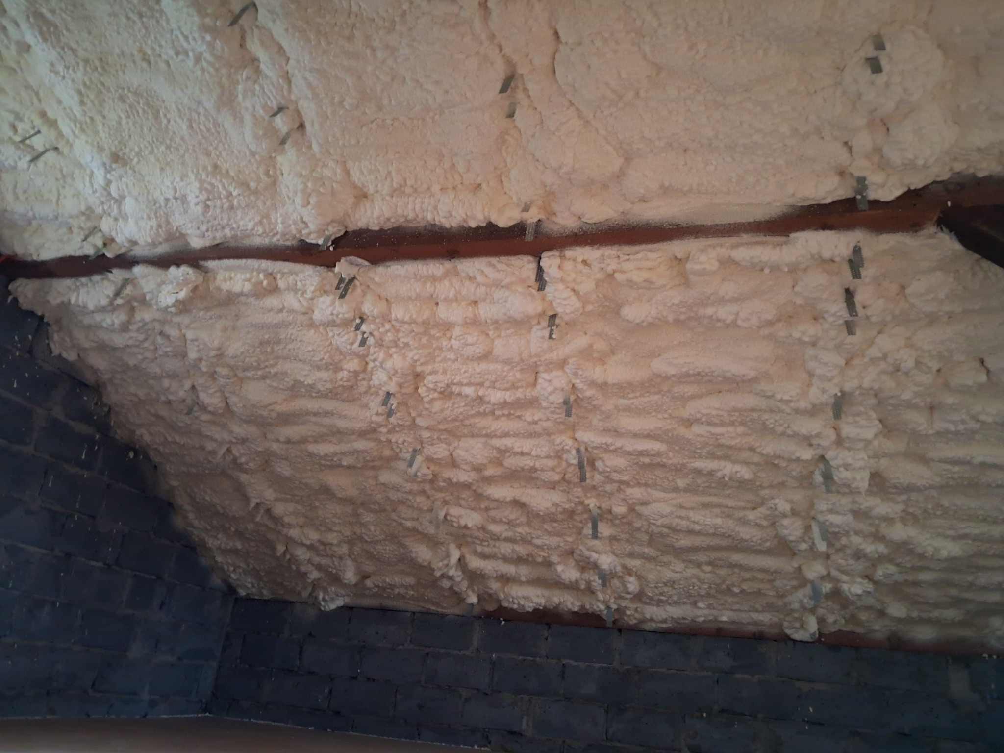 Poddasze z widoczną warstwą izolacji termicznej z pianki poliuretanowej, przymocowanej do krokwi za pomocą metalowych klamer, oraz fragmentem muru z ciemnych cegieł.