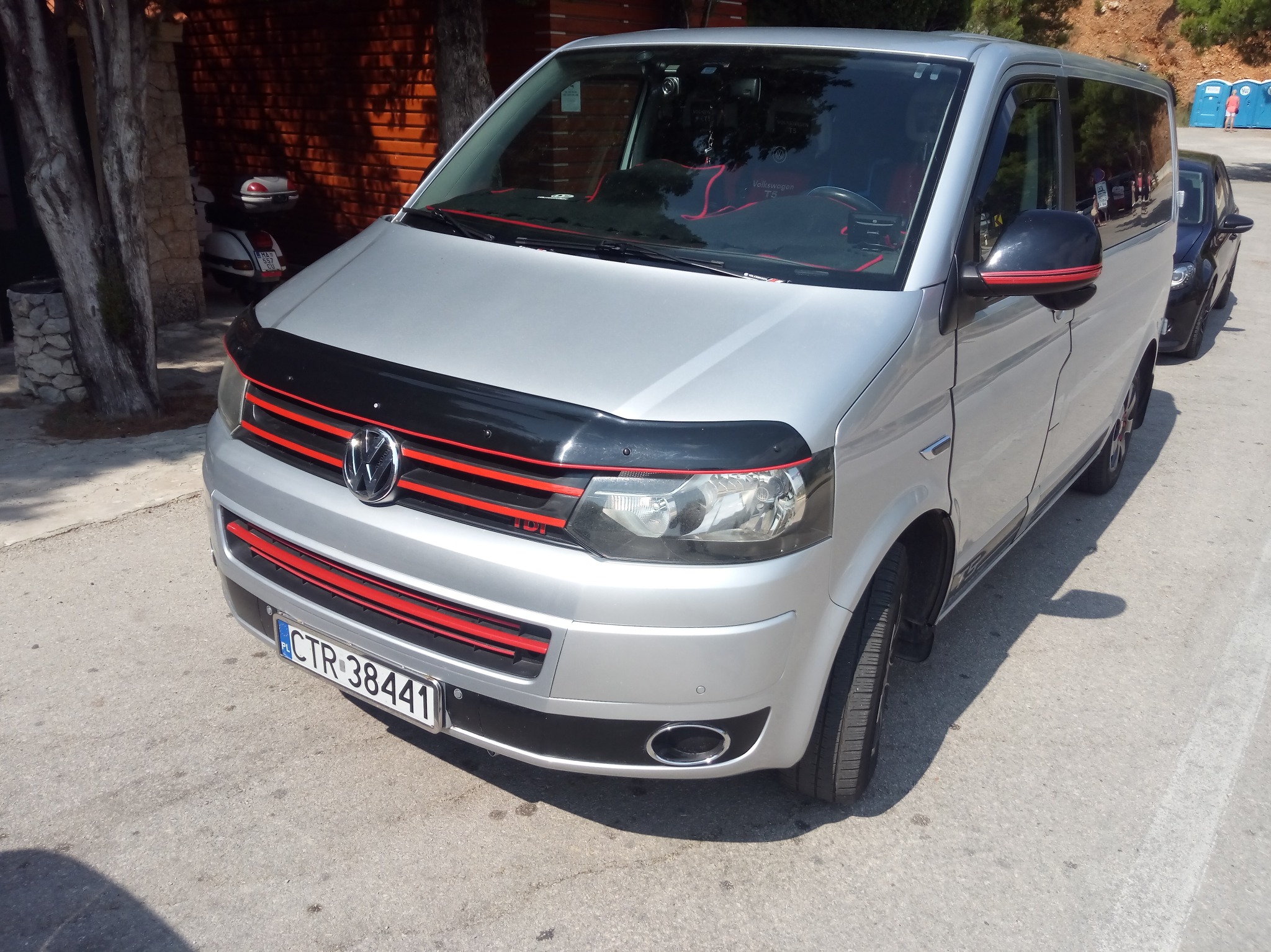 Srebrny Volkswagen Transporter T5 z czerwonymi akcentami, zaparkowany w Czarnowie na tle drzew i ceglanej ściany, z widocznym skuterem w tle.