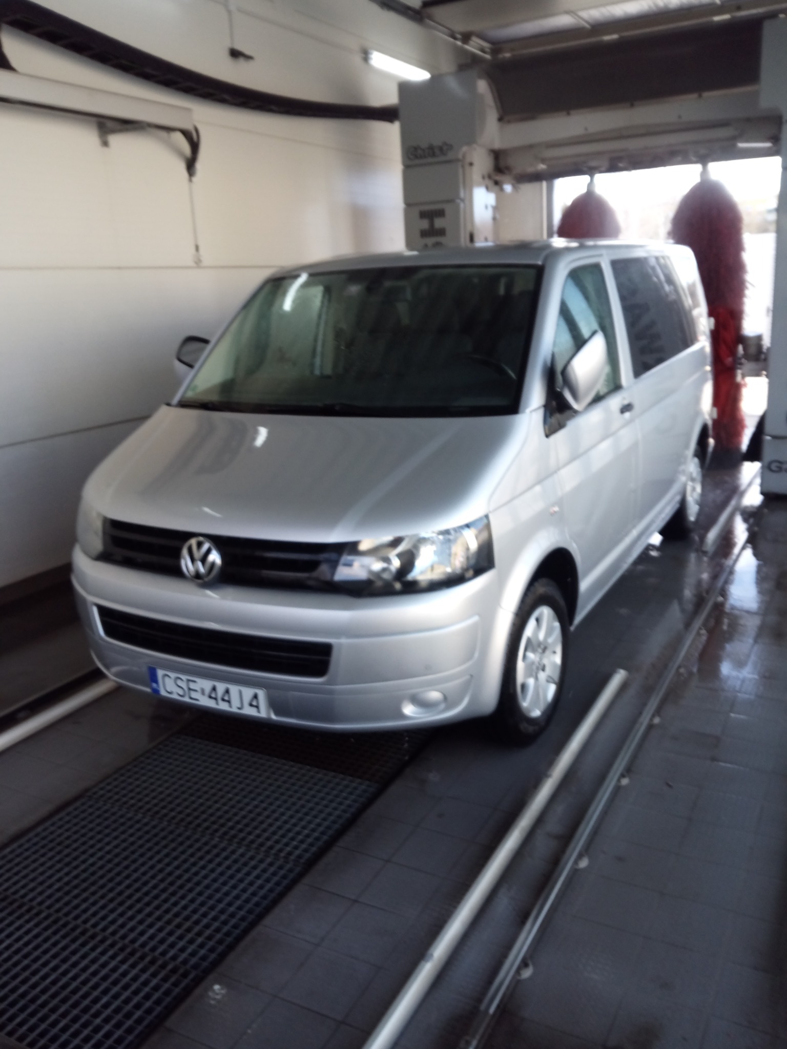 Srebrny Volkswagen Transporter w automatycznej myjni samochodowej, widoczne czerwone szczotki myjące i mokra posadzka.