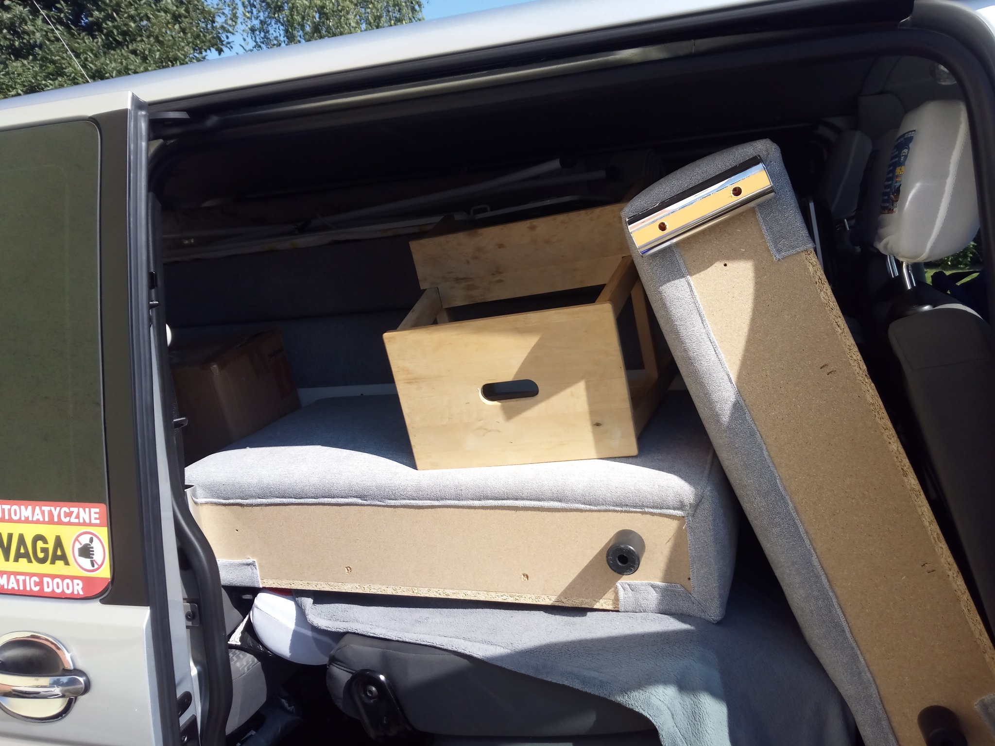 Wnętrze srebrnego busa z otwartymi drzwiami bocznymi, załadowane drewnianymi skrzynkami, tapicerowanymi elementami meblowymi i kartonem, widoczna naklejka ostrzegawcza na drzwiach.