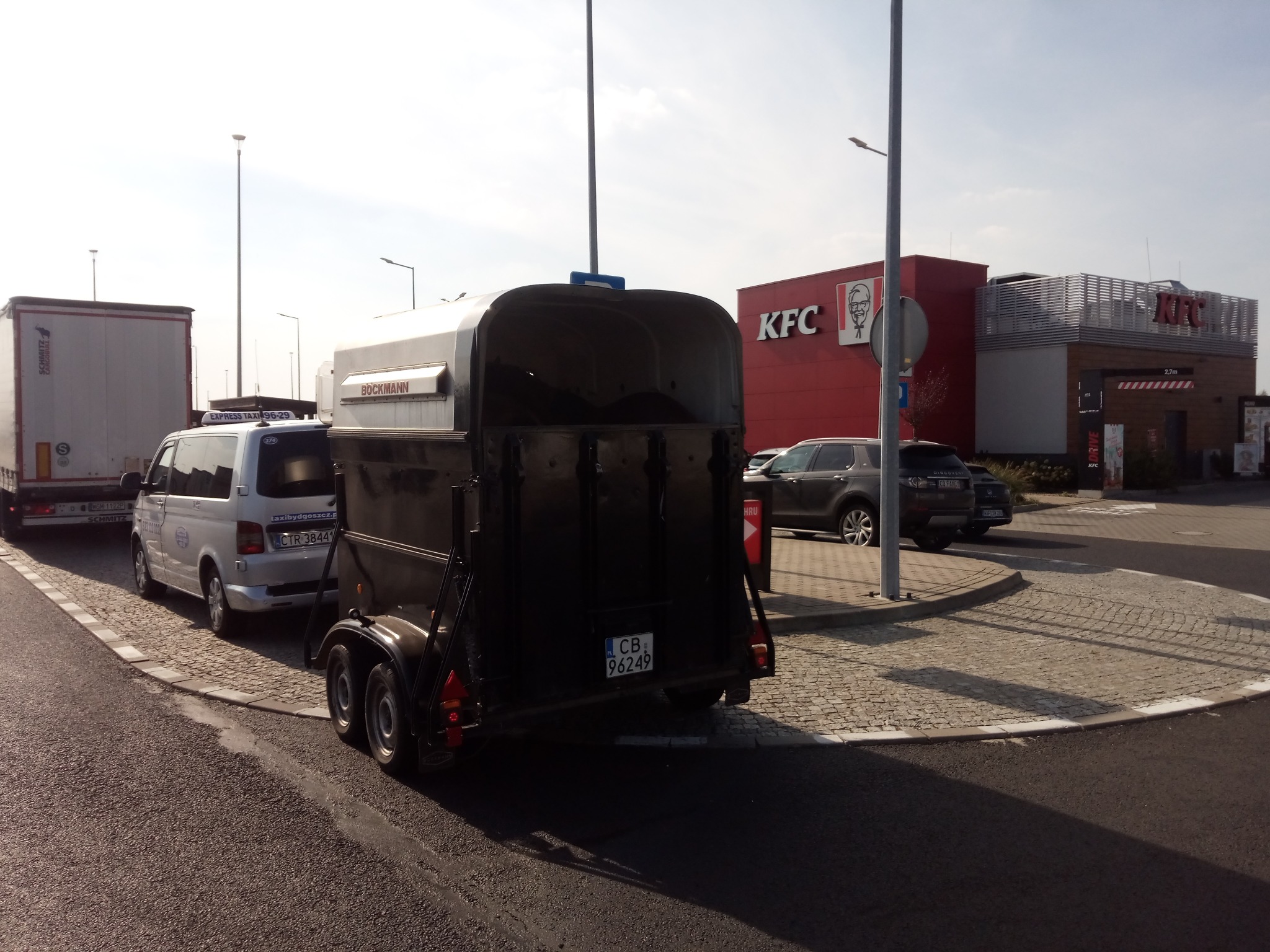 Przyczepa do transportu koni marki Böckmann zaparkowana na parkingu obok białego busa i ciężarówki, w tle budynek KFC.