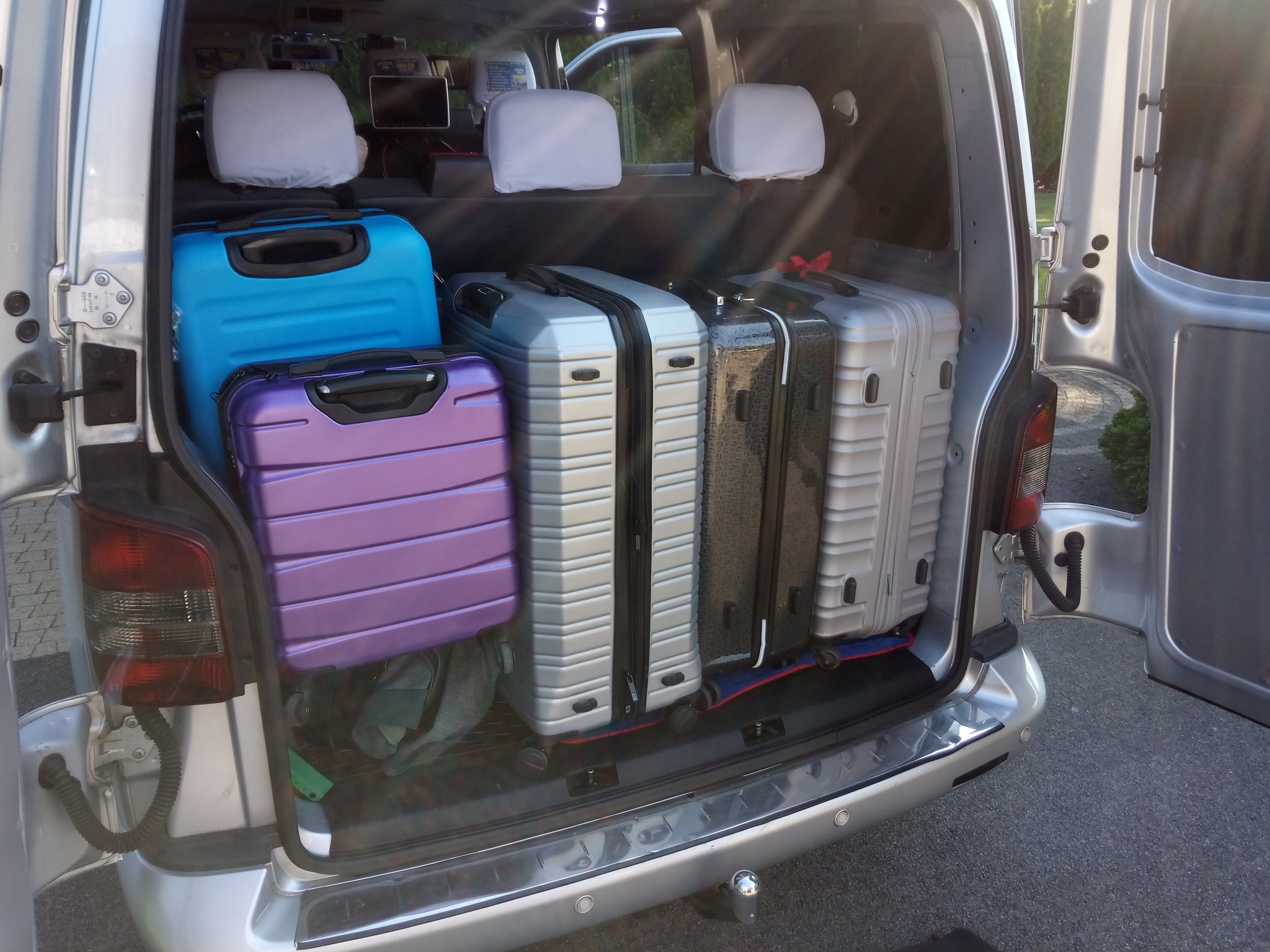Bagażnik srebrnego vana wypełniony kolorowymi walizkami: niebieską, fioletową, srebrną i czarną. Widoczne zagłówki foteli i otwarte drzwi bagażnika.