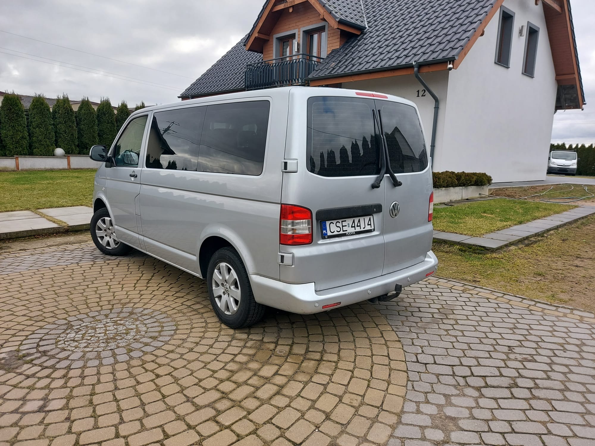 Srebrny bus Volkswagen Transporter zaparkowany na brukowanym podjeździe przed domem z szarą dachówką, widok z tyłu.