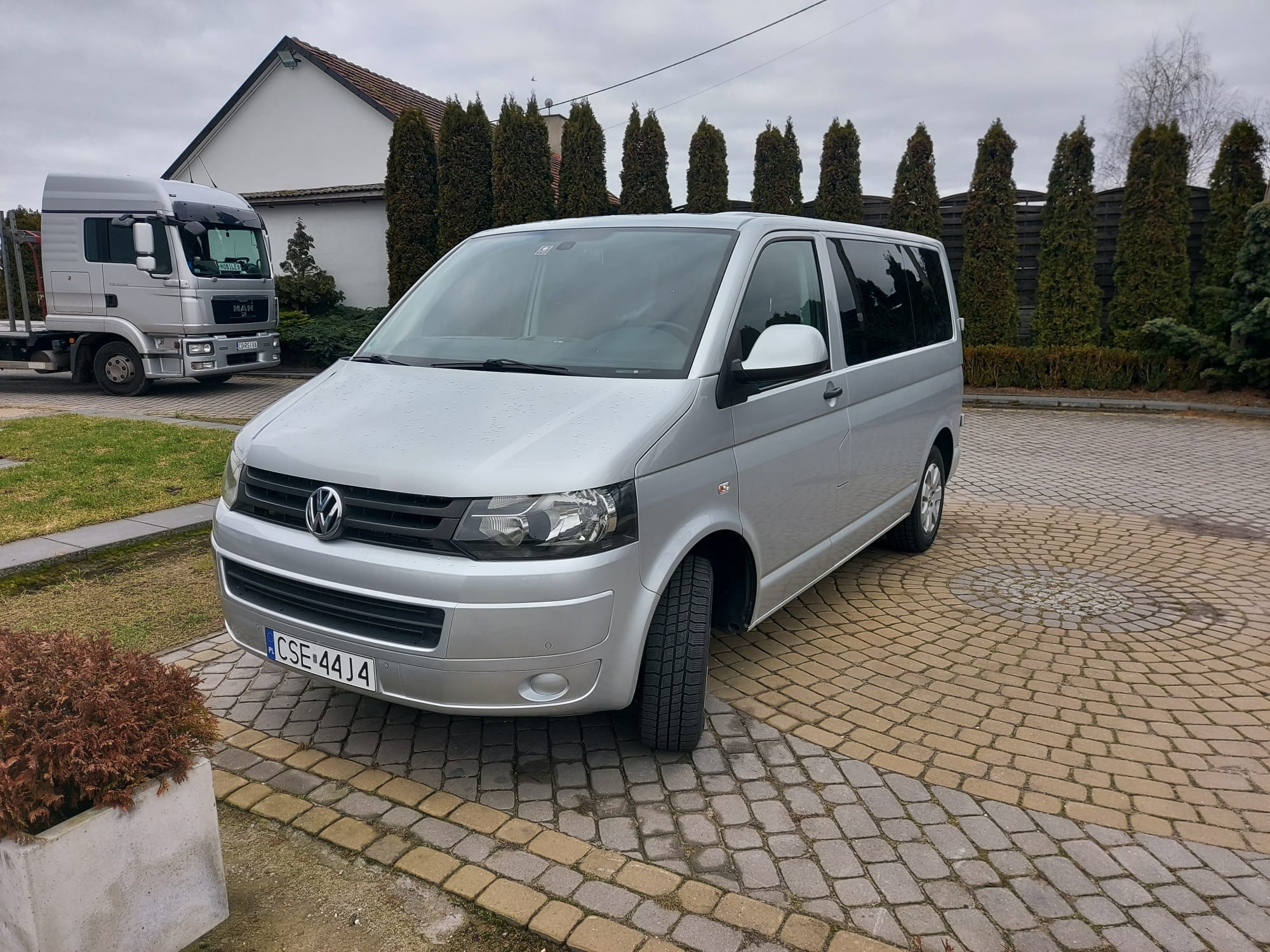 Srebrny Volkswagen Transporter T5 zaparkowany na kostce brukowej, z widoczną tablicą rejestracyjną CSE 44J4 i ciężarówką MAN w tle.