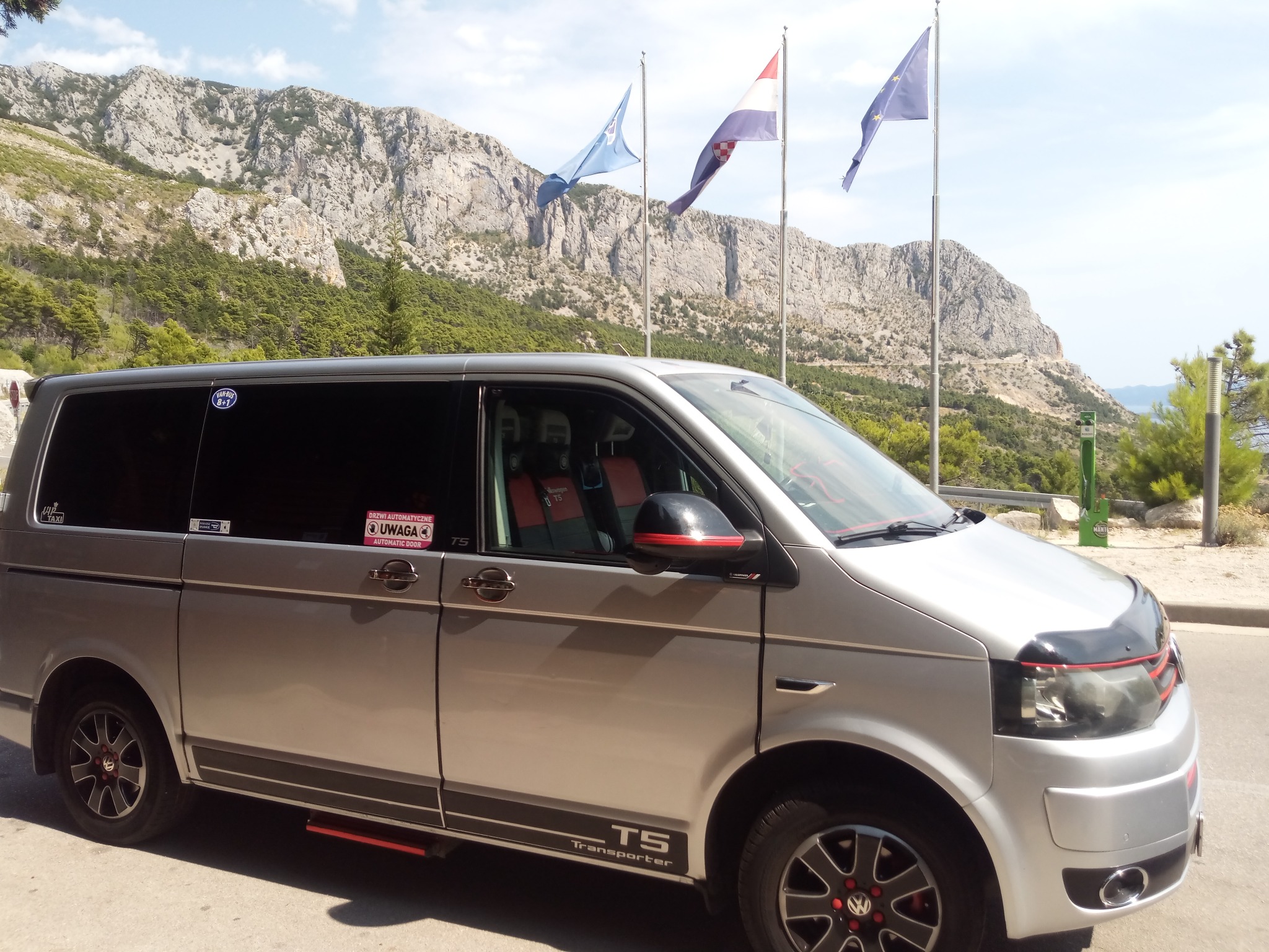 Srebrny bus Volkswagen Transporter T5 z naklejkami informacyjnymi na szybach, zaparkowany na tle górskiego krajobrazu z powiewającymi flagami Chorwacji i Unii Europejskiej.
