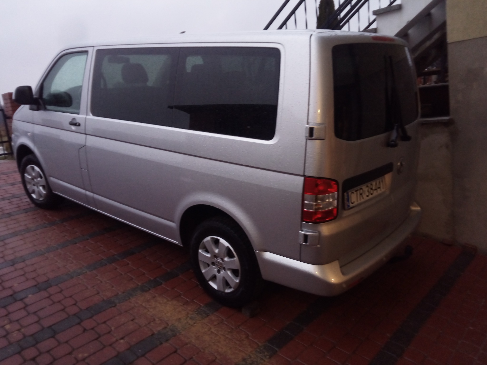 Srebrny van Volkswagen Transporter T5 zaparkowany na czerwonej kostce brukowej w pochmurny dzień, widok z tyłu z lekkim kątem.