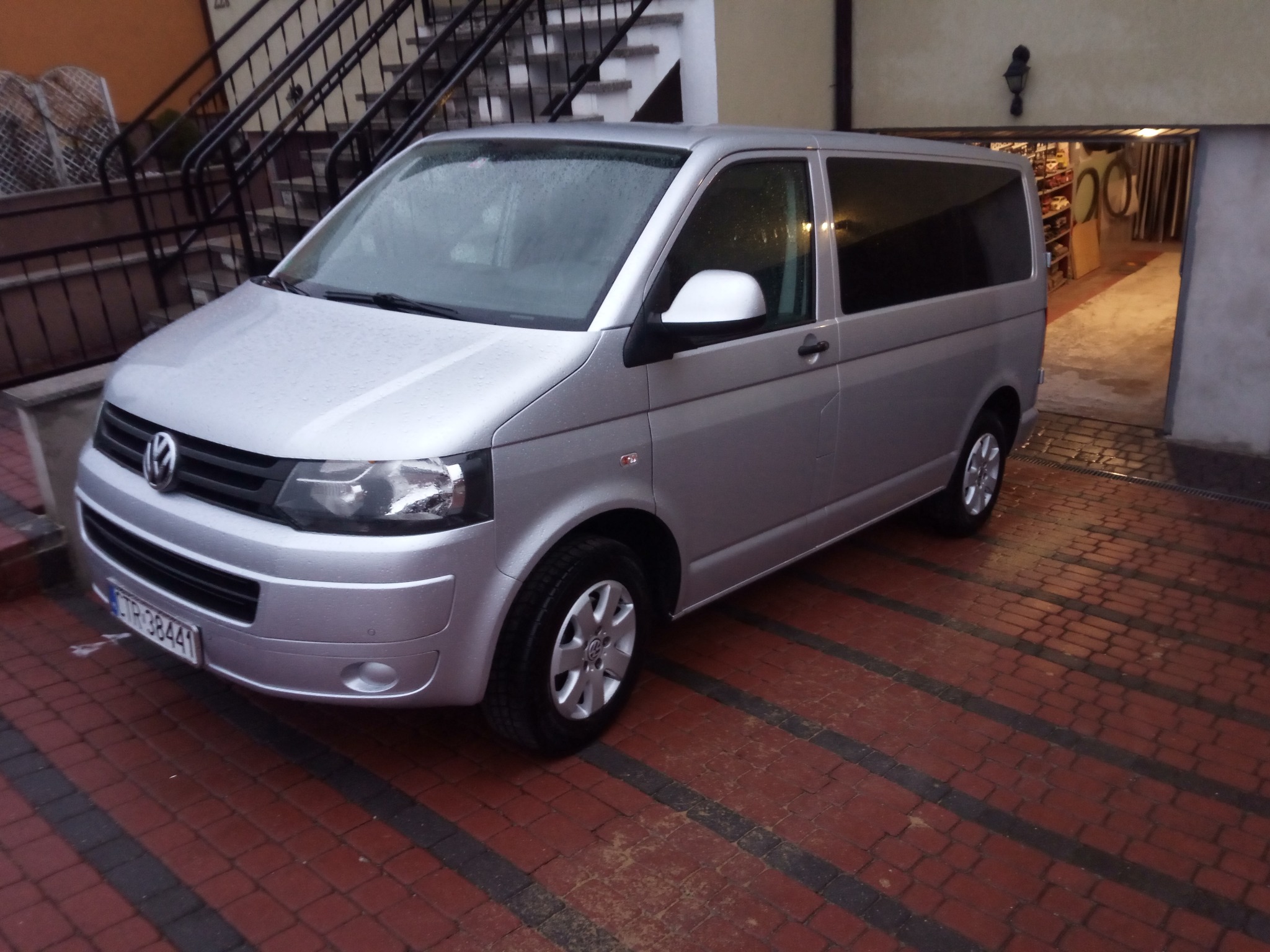 Srebrny van Volkswagen Transporter zaparkowany na czerwono-czarnym brukowanym podjeździe przed budynkiem z czarną balustradą schodów i otwartym garażem.