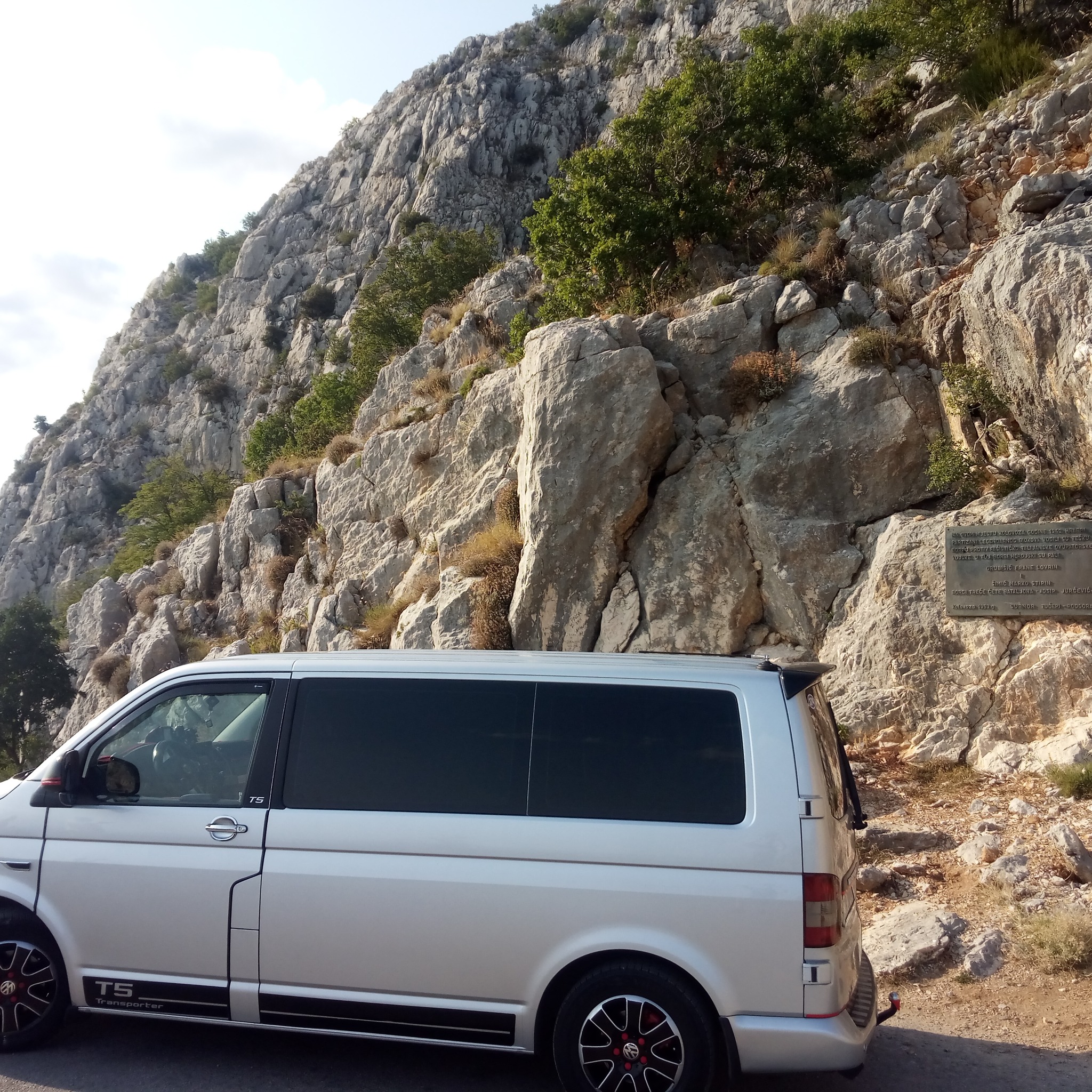 Srebrny bus Transporter T5 zaparkowany na tle skalistej góry z tablicą pamiątkową, widok z boku, czarne szyby
