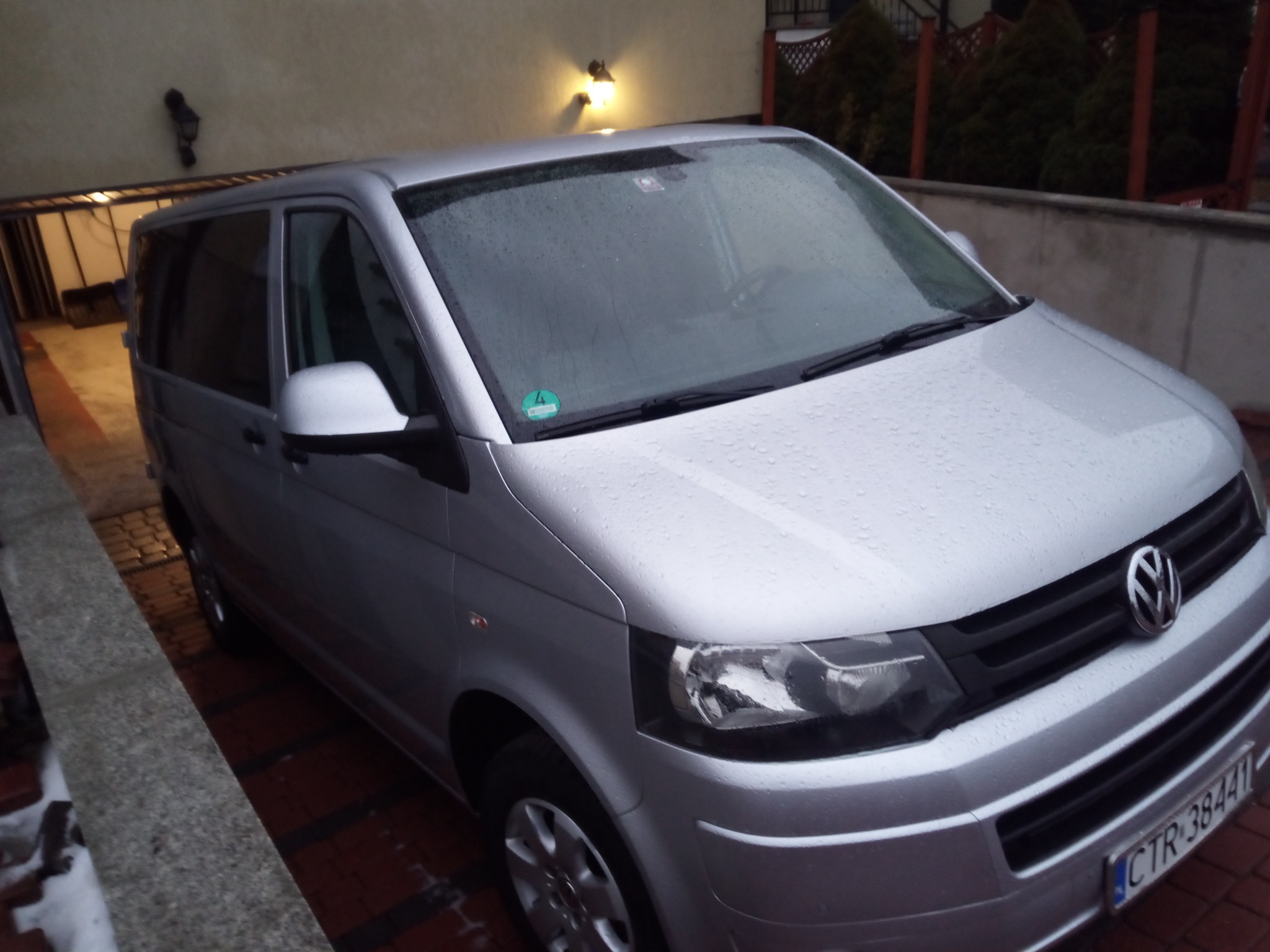 Srebrny Volkswagen Transporter T5, mokry od deszczu, zaparkowany na posesji z czerwonej kostki brukowej, widoczny wjazd do garażu i oświetlenie zewnętrzne.