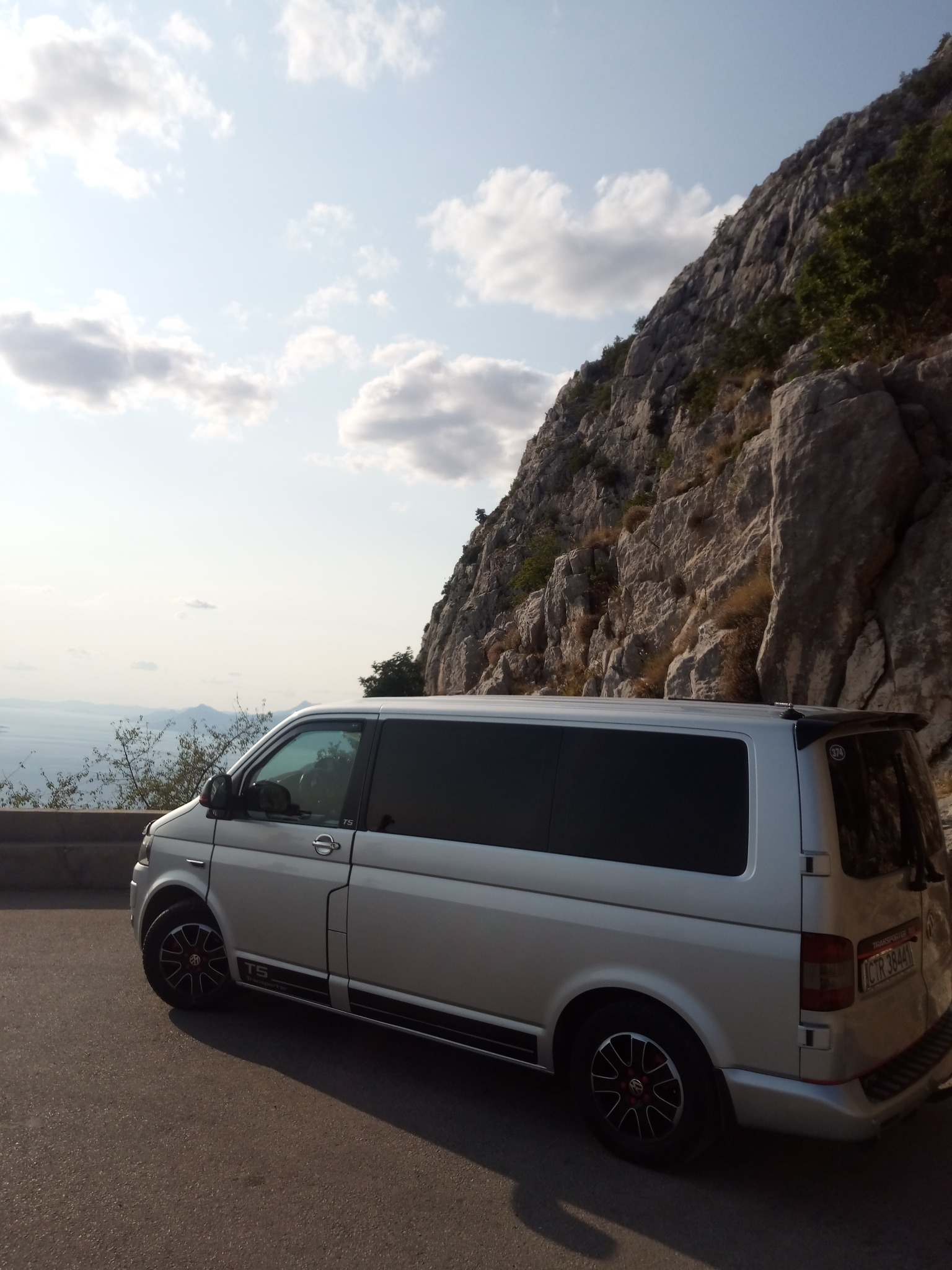 Srebrny bus T5 z przyciemnianymi szybami i czarnymi felgami z czerwonymi akcentami, zaparkowany na tle skalistej góry i widoku na morze w oddali.