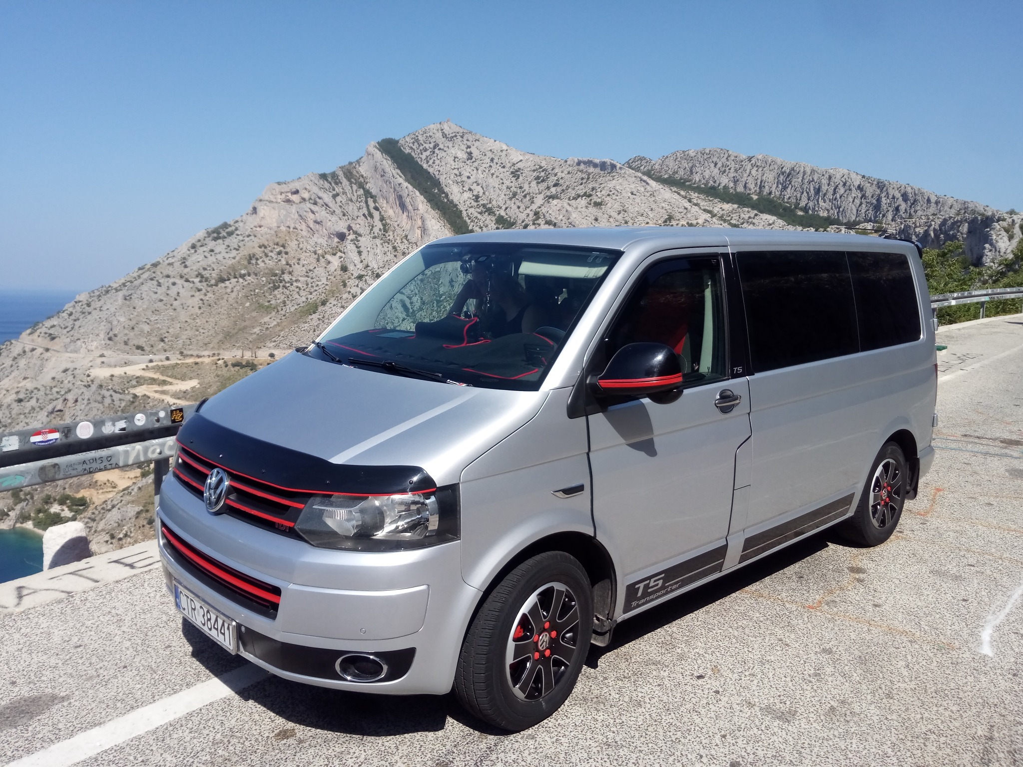 Srebrny Volkswagen Transporter T5 na tle skalistego krajobrazu z widokiem na morze, z czarnym deflektorem na masce i czerwonymi akcentami na grillu i felgach.