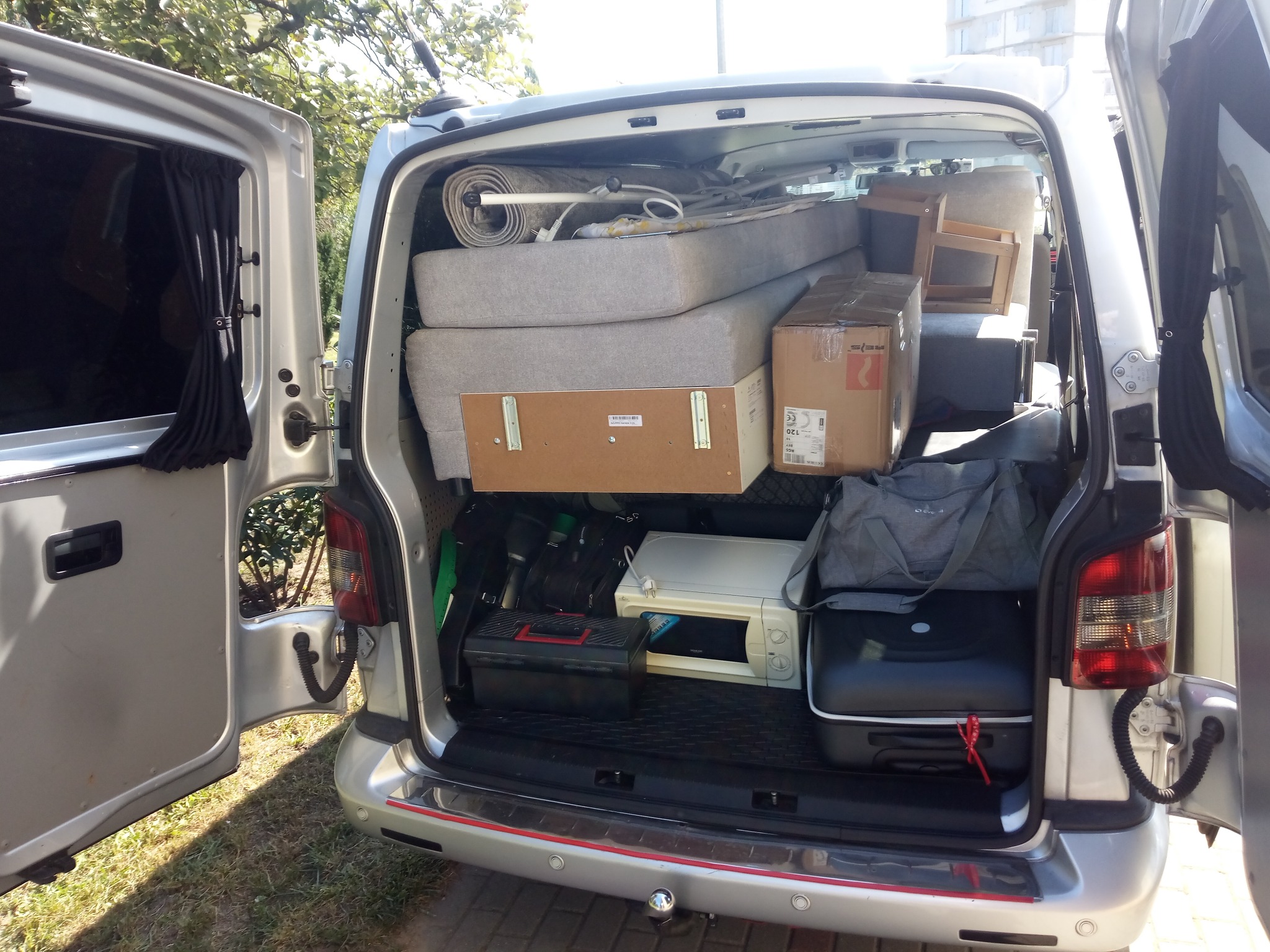 Srebrny van z otwartymi tylnymi drzwiami, załadowany meblami, kartonami i sprzętem AGD podczas przeprowadzki, widoczne zasłony w oknie.