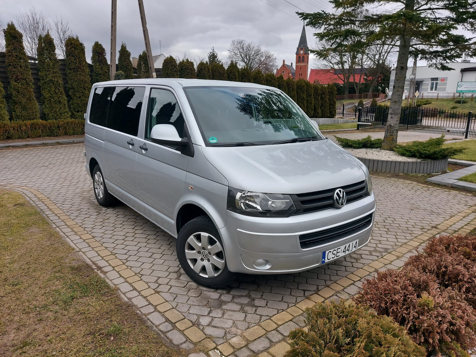 Srebrny Volkswagen Transporter T5 zaparkowany na kostce brukowej przed zadbanym ogrodem z widokiem na ceglany kościół w tle.