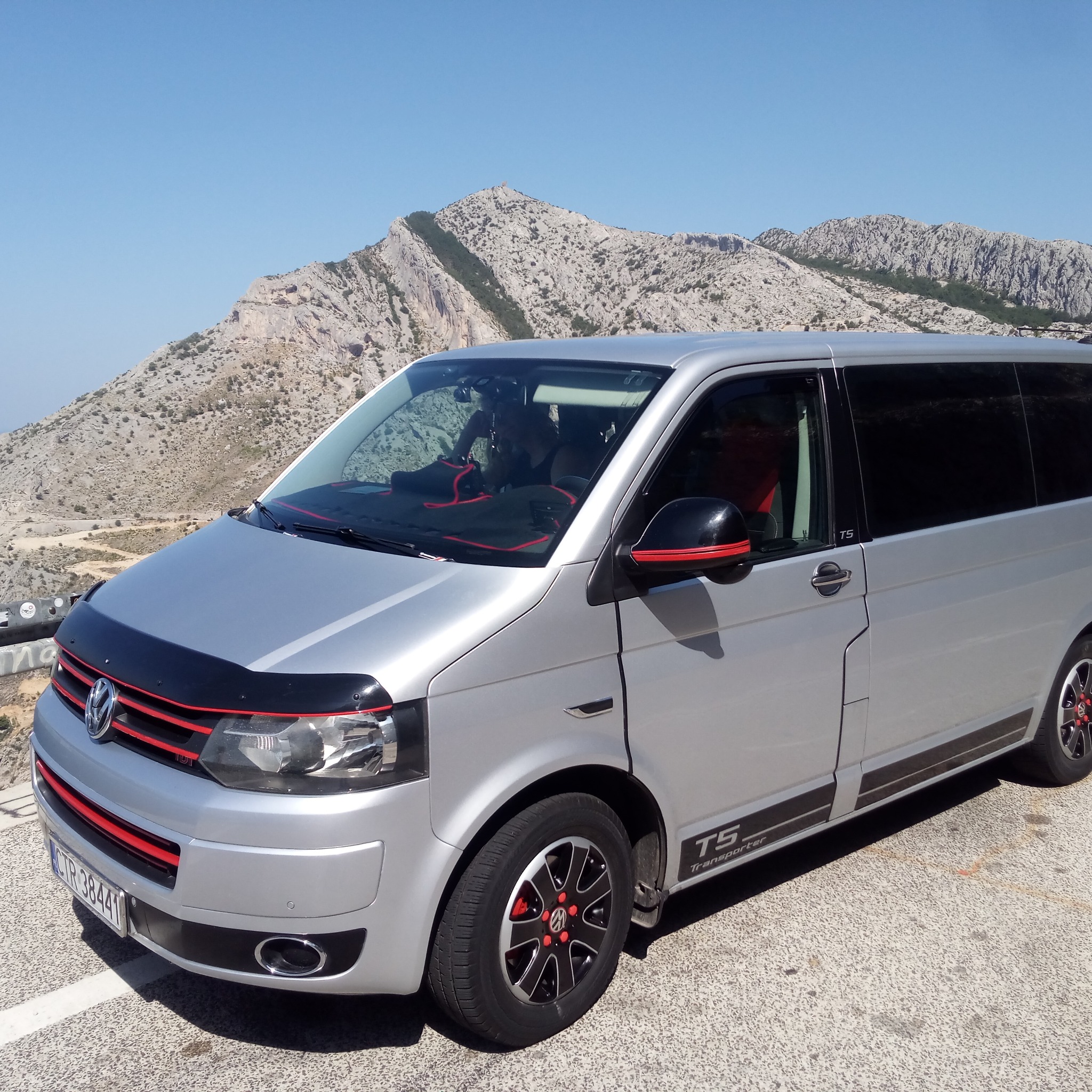 Srebrny Volkswagen T5 Transporter z czerwonymi akcentami na tle górskiego krajobrazu w słoneczny dzień.