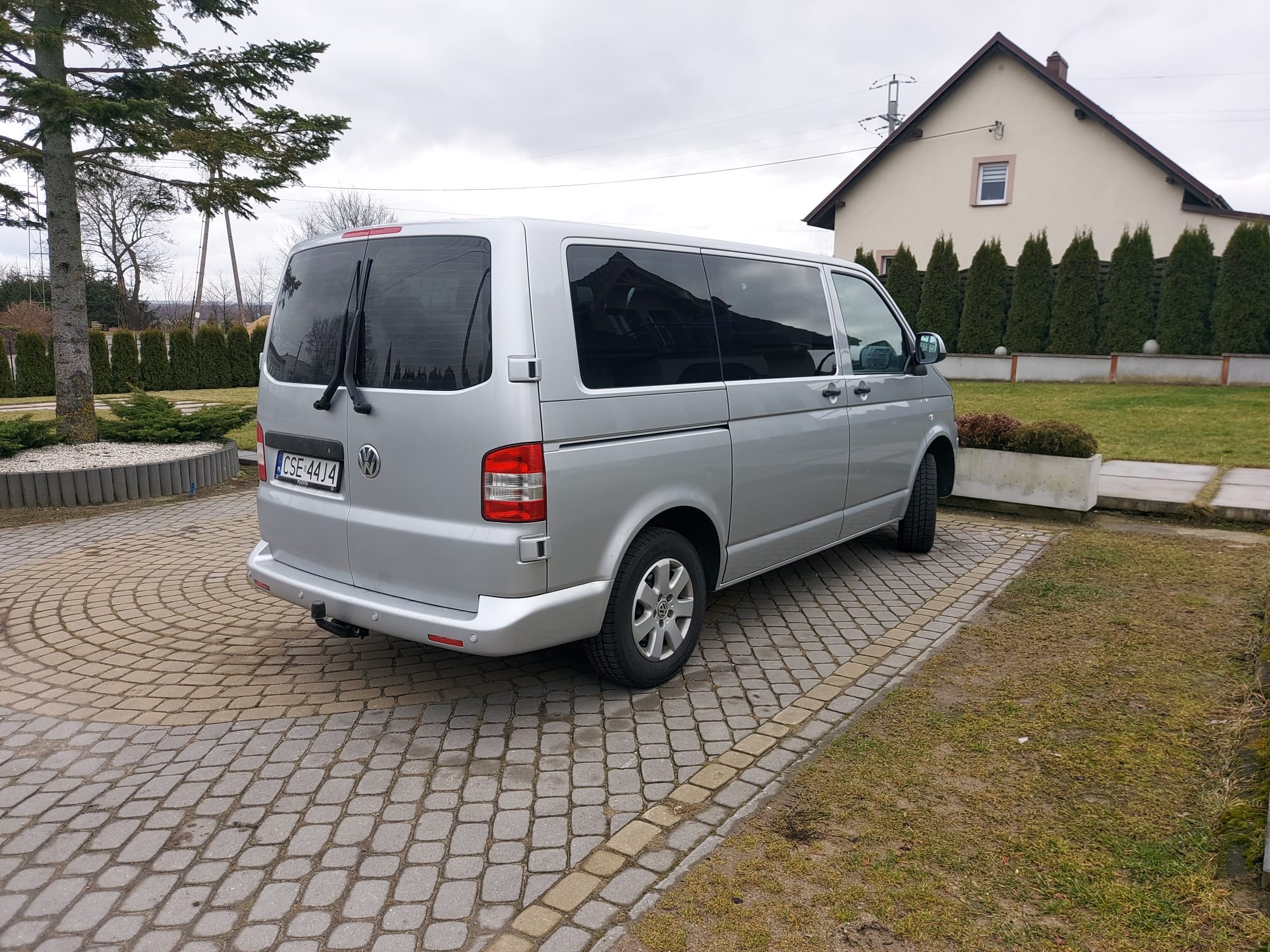 Srebrny bus Volkswagen Transporter zaparkowany na brukowanym podjeździe przed domem z zadbanym ogrodem i żywopłotem.