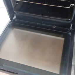 Monika Winiarska - Wnętrze brudnego piekarnika z widocznymi przypaleniami na ruszcie i tłustą szybą, otwarte drzwiczki ukazują drewnianą podłogę.