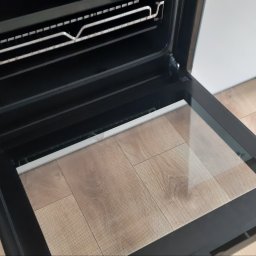 Monika Winiarska - Wnętrze piekarnika elektrycznego z otwartymi drzwiczkami, widoczna płyta grzejna i ruszt, na podłodze jasne panele.