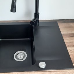 Monika Winiarska - Czarny, kwadratowy zlew granitowy z chromowanym odpływem i czarną baterią, zamontowany w blacie imitującym drewno, z widocznym gniazdkiem elektrycznym na białej ścianie w tle.