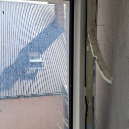 Monika Winiarska - Widok z brudnego okna na blaszany dach z kominem i okno dachowe, fragment ściany z resztkami taśmy malarskiej.