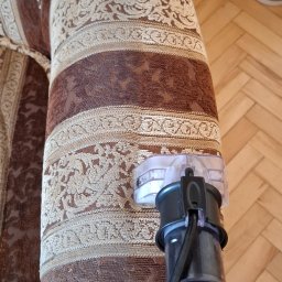 Monika Winiarska - Czyszczenie tapicerki kanapy z brązowym wzorem za pomocą odkurzacza piorącego, widoczna dysza ssąca i fragment parkietu w tle.