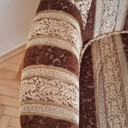 Monika Winiarska - Fragment brązowo-beżowej kanapy z dekoracyjnym wzorem, widoczny bok i oparcie, na podłodze parkiet.