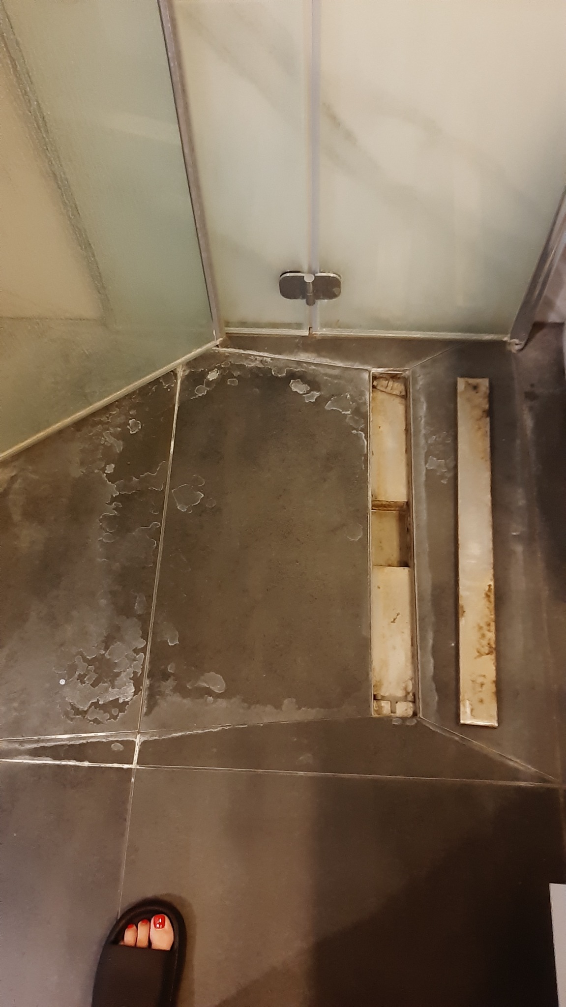 Zacieki i osad na ciemnych płytkach pod prysznicem, widoczny odpływ liniowy z rdzawym nalotem oraz fragment stopy w klapku z czerwonym lakierem na paznokciach.