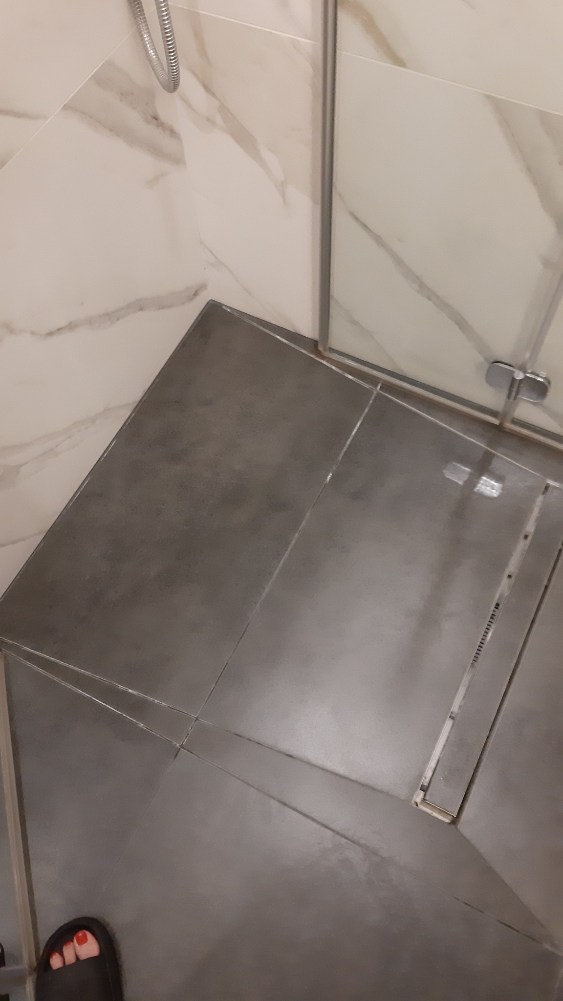 Szare płytki podłogowe w kabinie prysznicowej z odpływem liniowym, fragment stopy w klapku w lewym dolnym rogu, marmurowe płytki ścienne w tle.