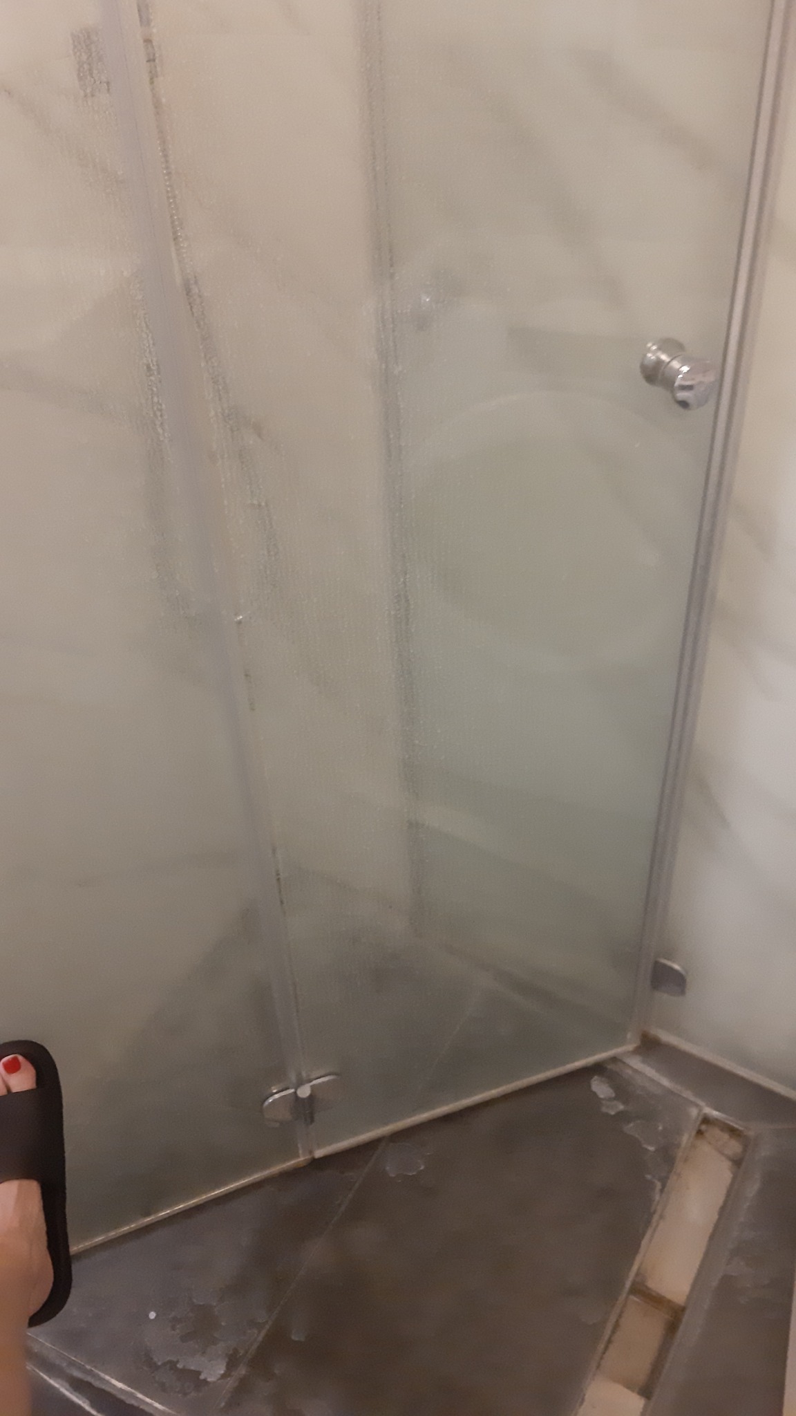 Brudna kabina prysznicowa z zaciekami na szkle i plamami na podłodze, widoczny fragment stopy w klapku z czerwonym paznokciem.