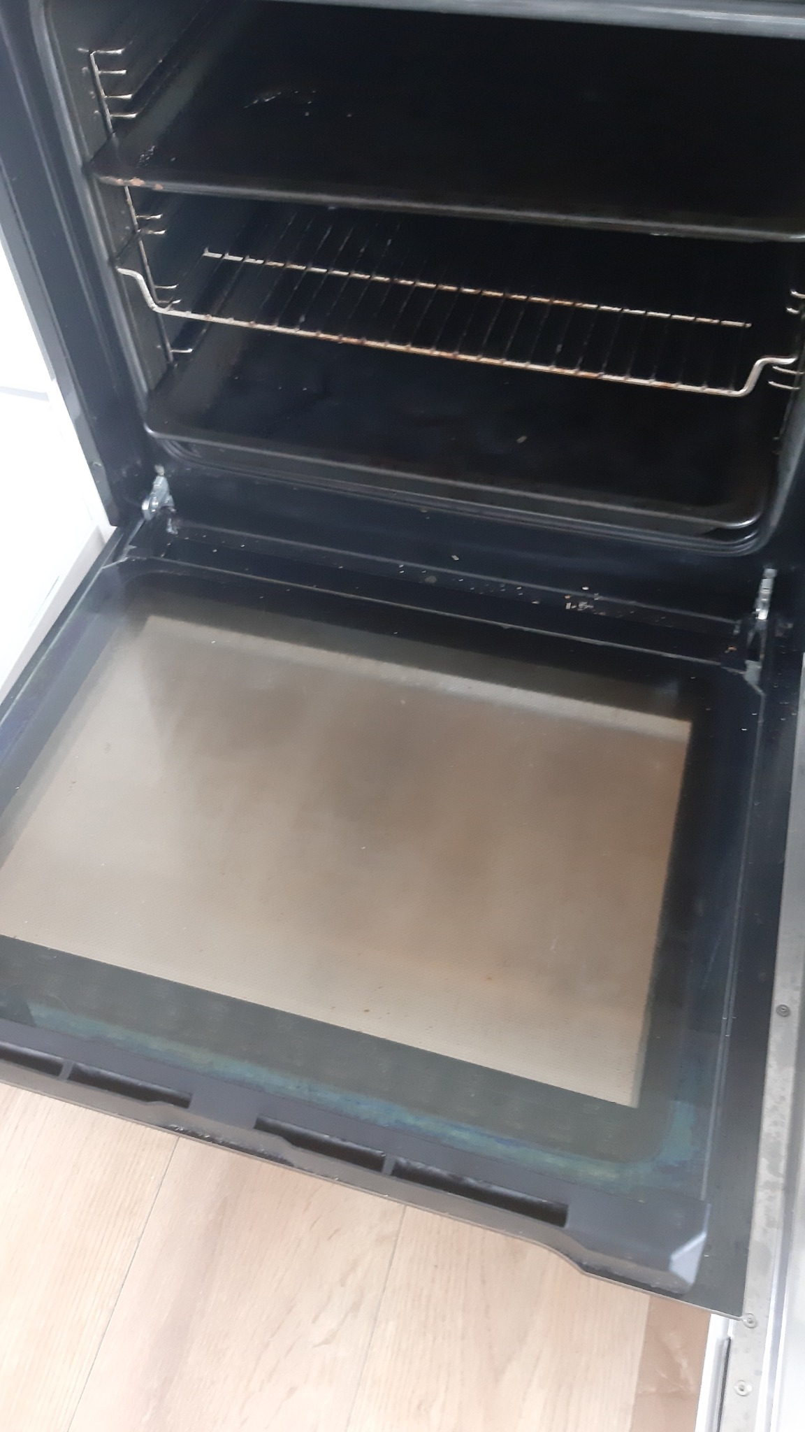 Wnętrze brudnego piekarnika z widocznymi przypaleniami na ruszcie i tłustą szybą, otwarte drzwiczki ukazują drewnianą podłogę.