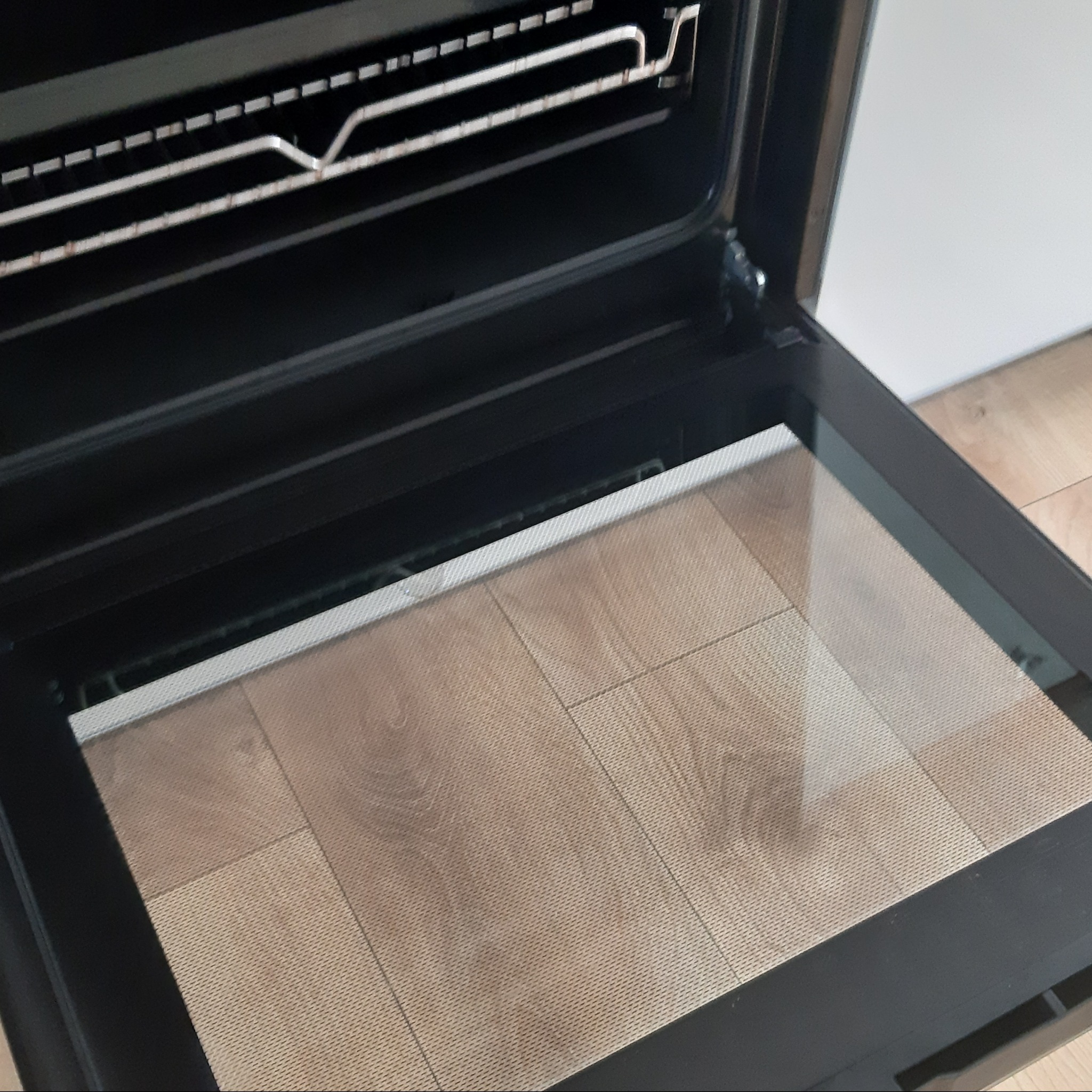 Wnętrze piekarnika elektrycznego z otwartymi drzwiczkami, widoczna płyta grzejna i ruszt, na podłodze jasne panele.