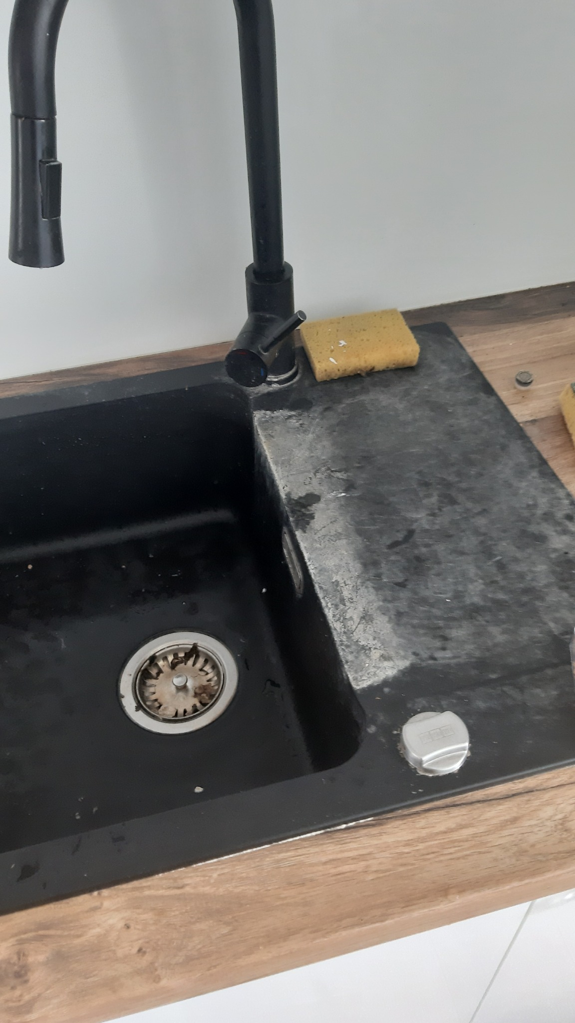 Czarny, brudny zlew granitowy z resztkami jedzenia w odpływie, obok leży gąbka i metalowe elementy, widoczne ślady po remoncie.