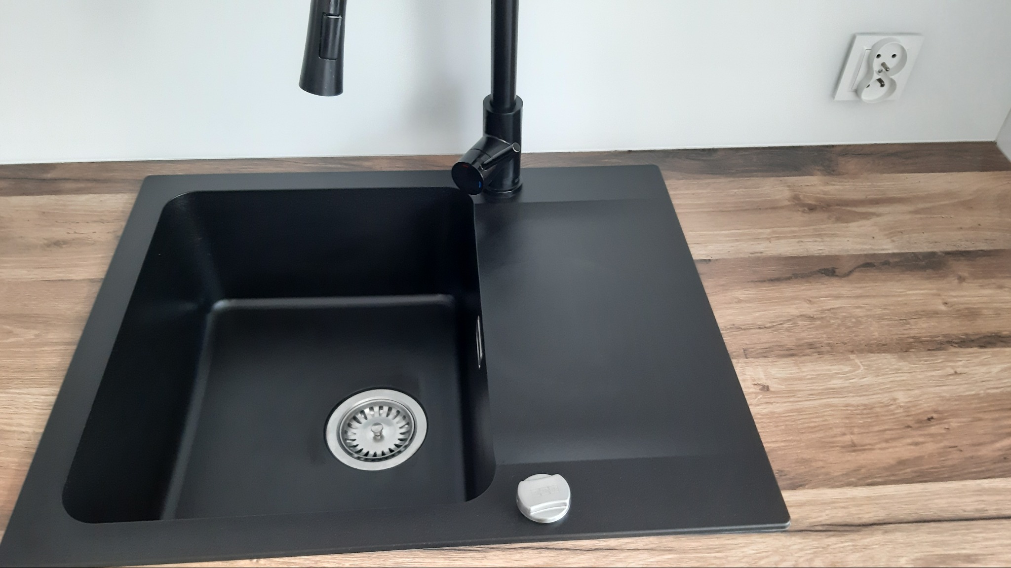 Czarny, kwadratowy zlew granitowy z chromowanym odpływem i czarną baterią, zamontowany w blacie imitującym drewno, z widocznym gniazdkiem elektrycznym na białej ścianie w tle.