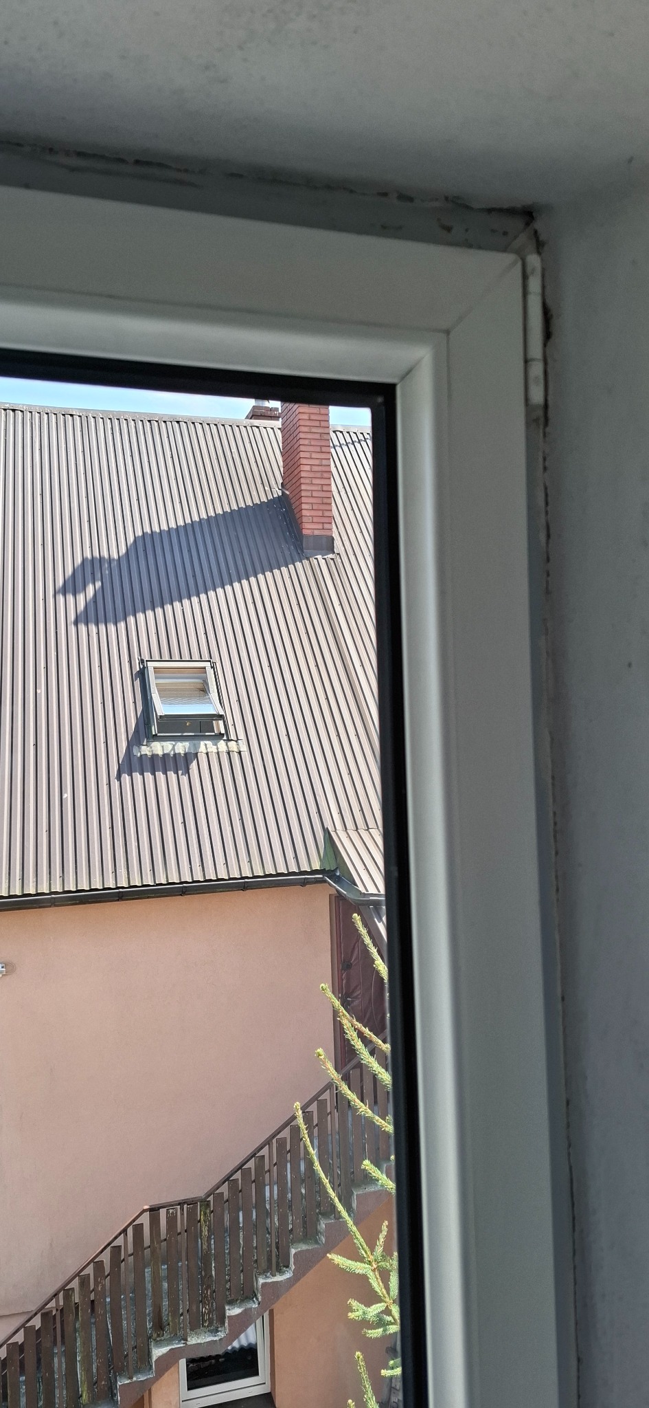 Widok z okna na zewnątrz, widoczny dach z blachy, komin i fragment budynku z drewnianymi schodami. Rama okienna z widocznymi zabrudzeniami.