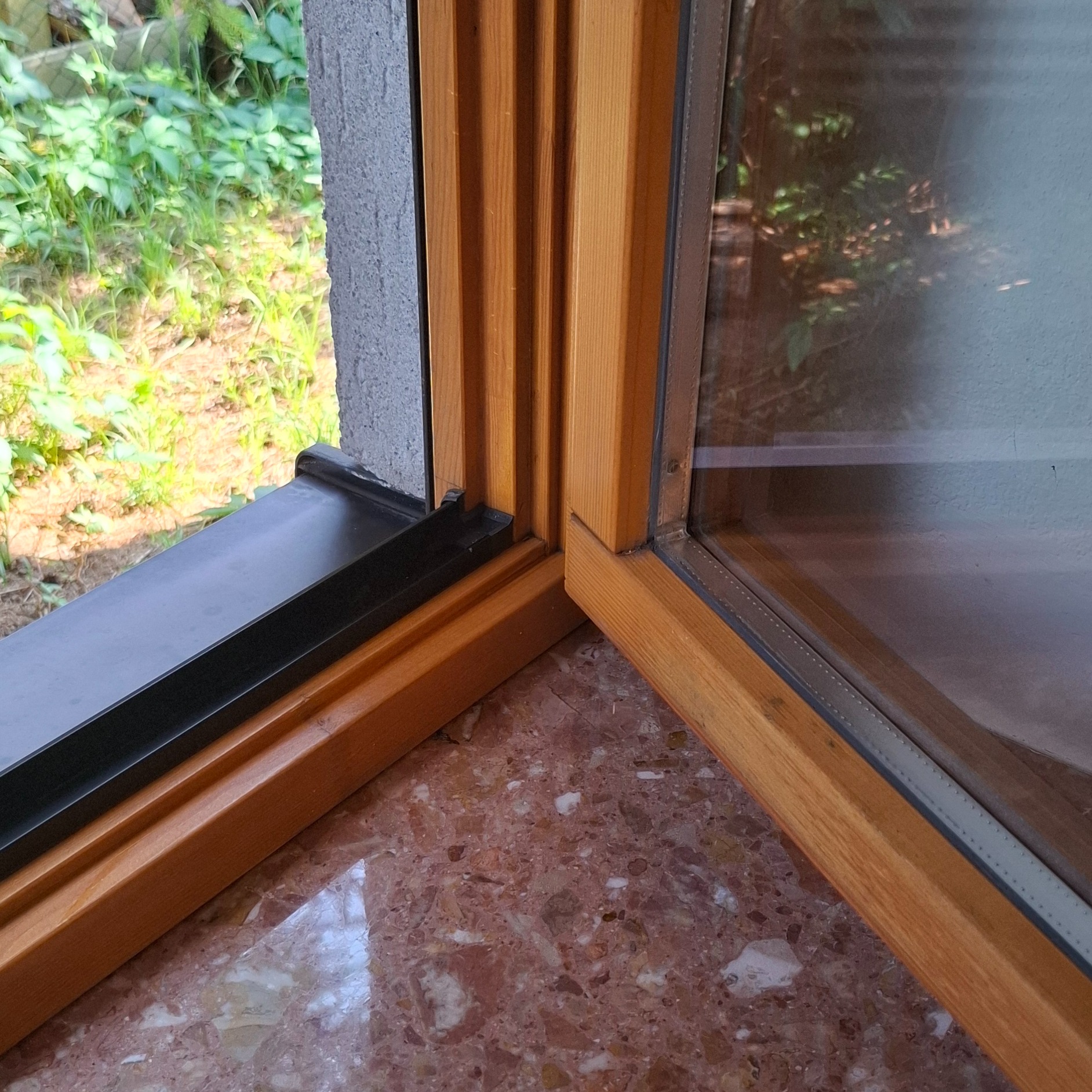 Fragment okna z drewnianą ramą i czarnym parapetem wewnętrznym na tle marmurowego blatu, widok na zieleń za oknem.