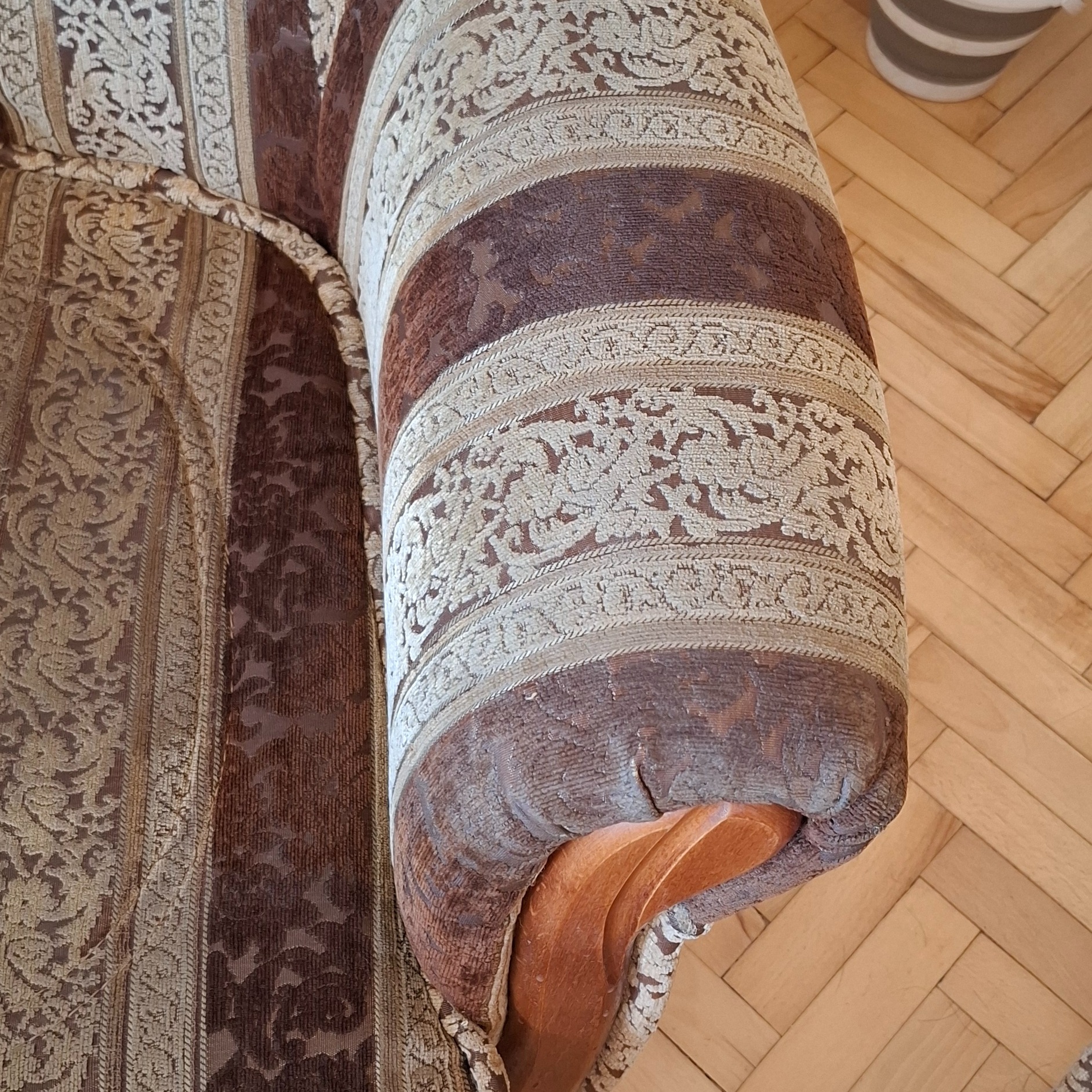 Fragment zniszczonego, brązowo-beżowego fotela z wzorzystą tapicerką, widoczny na tle parkietu; uszkodzone obicie na podłokietniku.
