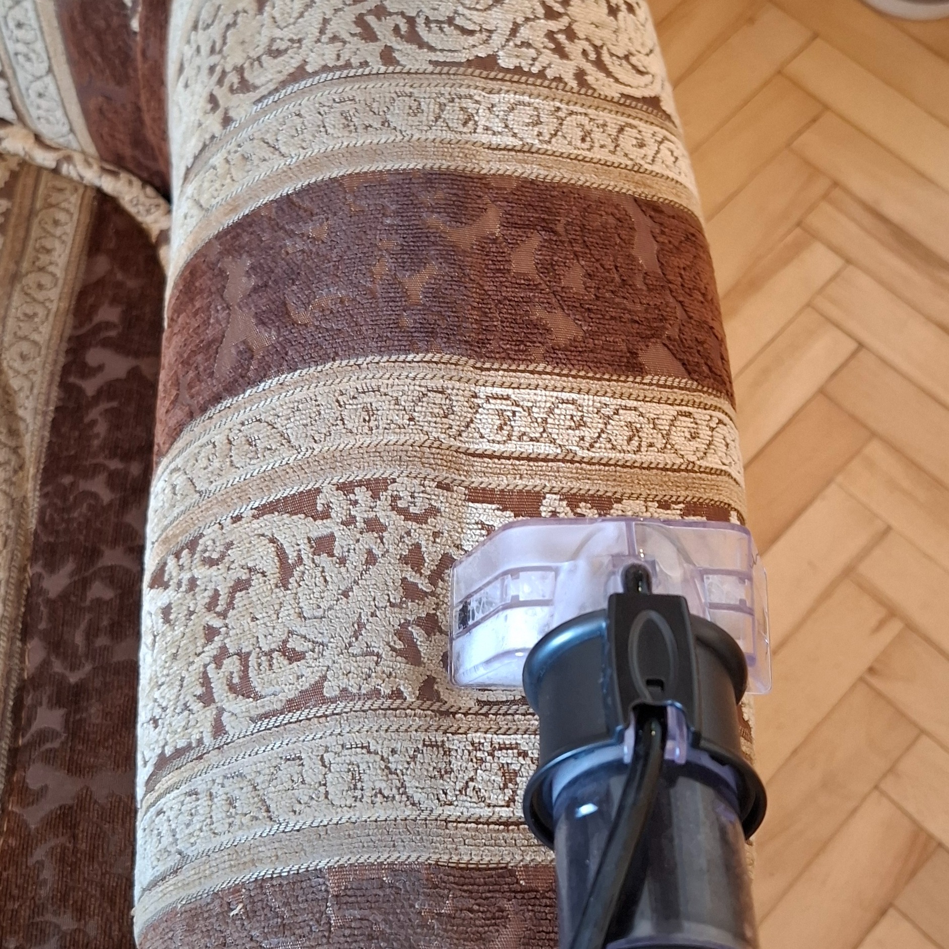 Czyszczenie tapicerki kanapy z brązowym wzorem za pomocą odkurzacza piorącego, widoczna dysza ssąca i fragment parkietu w tle.