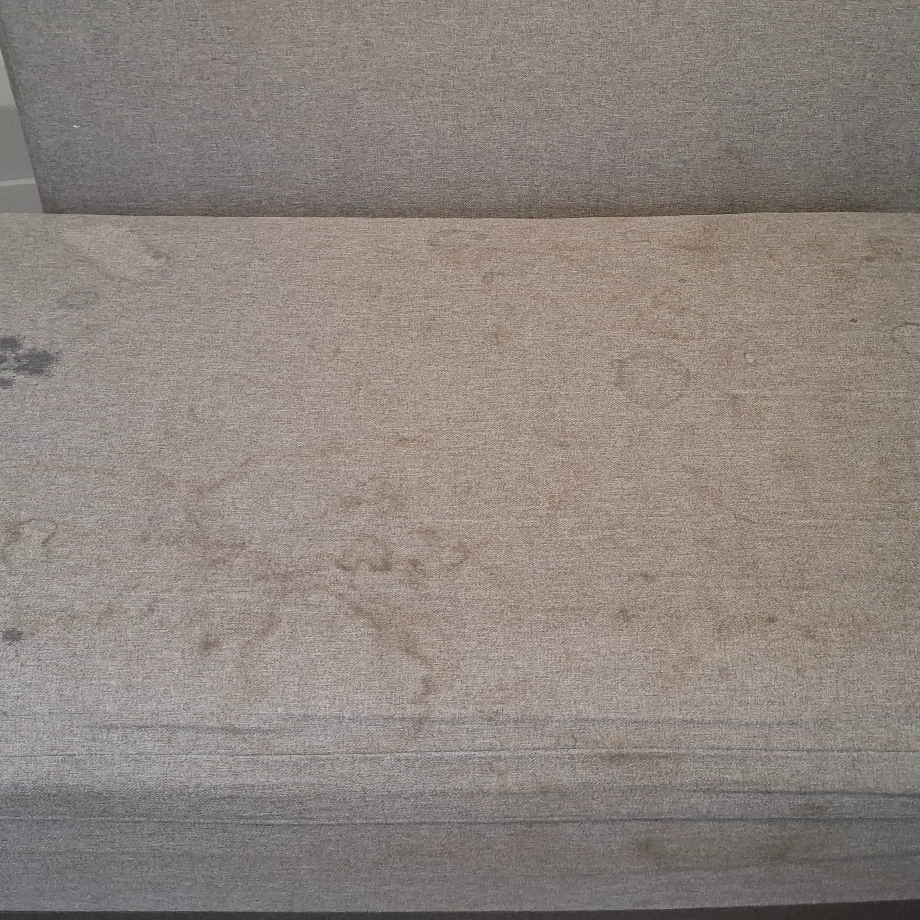 Beżowa tapicerka kanapy z widocznymi, ciemnymi plamami i okrągłymi zaciekami, wymagająca odświeżenia.